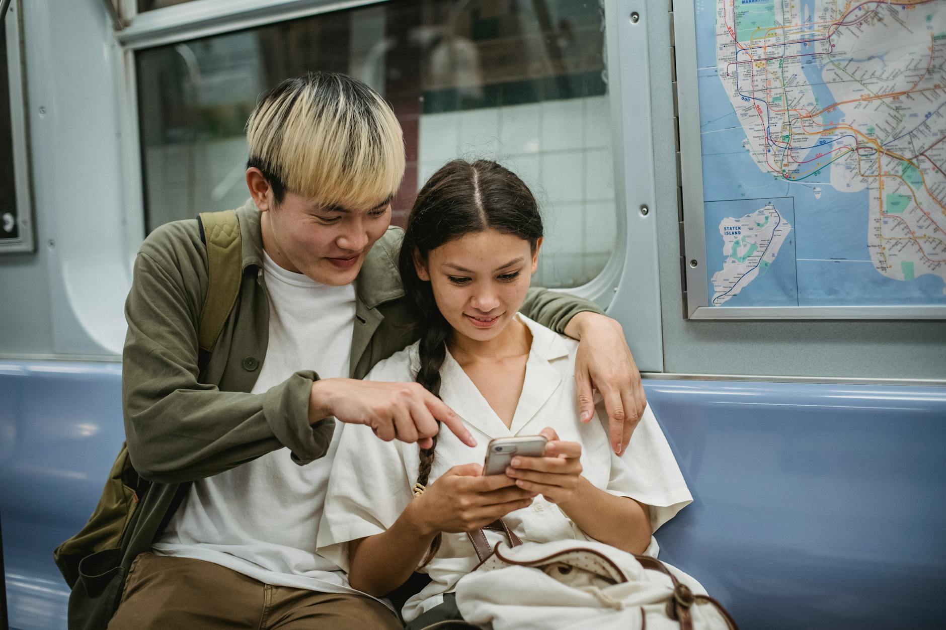 Happy young trendy Asian guy hugging smiling girlfriend and pointing at smartphone screen while commuting by subway train