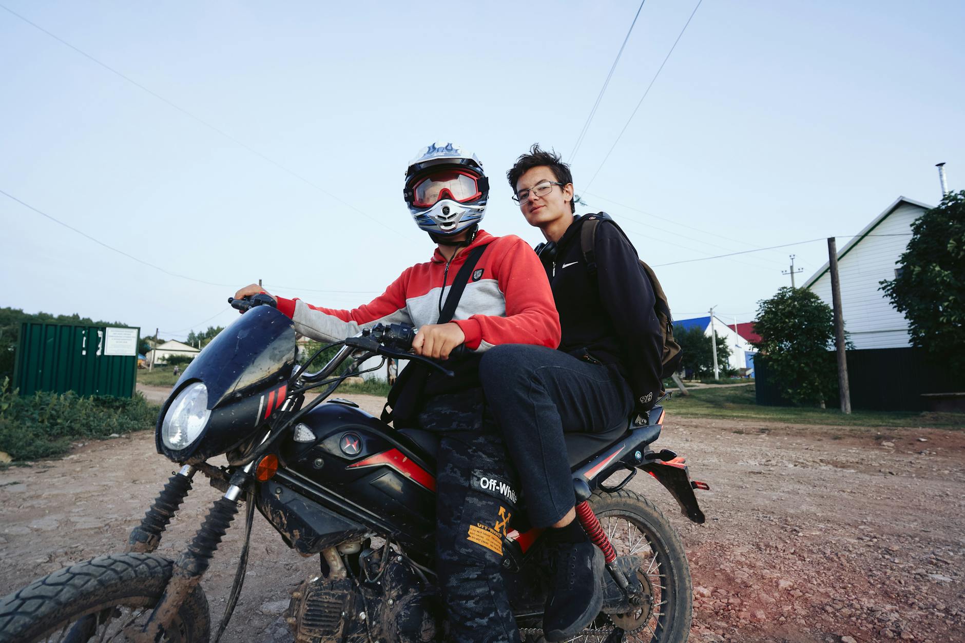 Two young adults on a dirt bike with safety gear on a rural road during daytime.