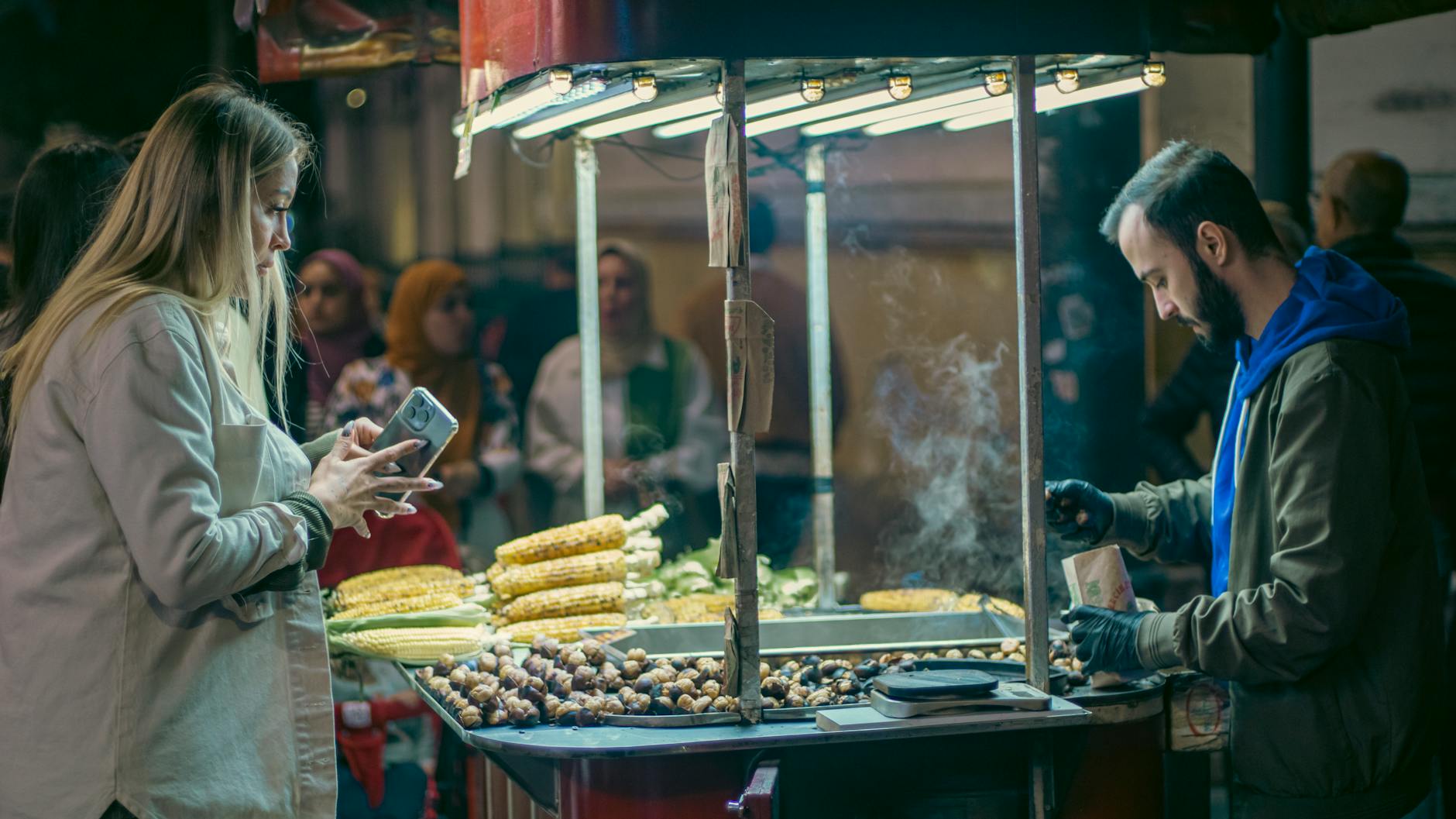 A lively street food market scene with a vendor selling corn and chestnuts to a diverse group of customers.