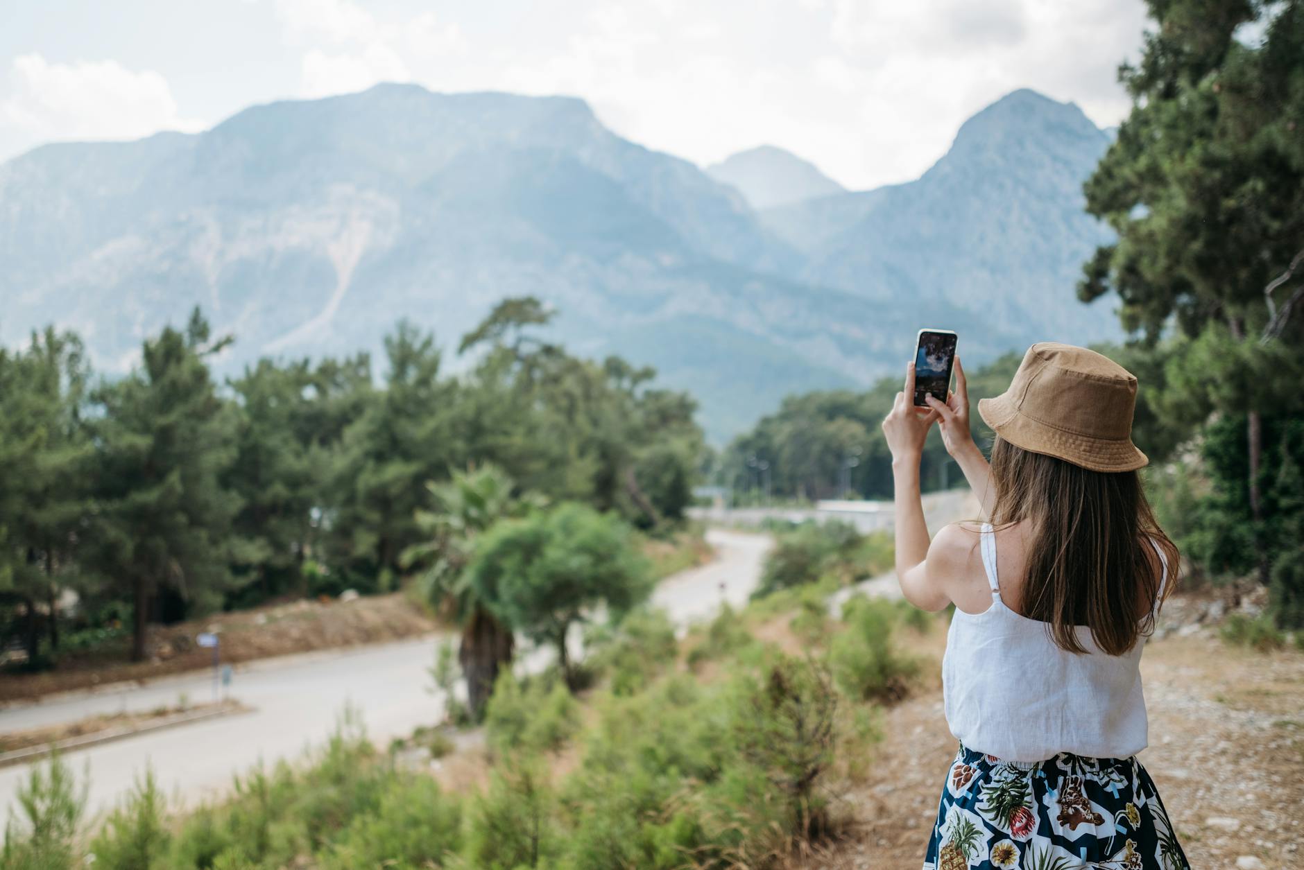 A woman in a hat photographs a mountain landscape with her phone during summer.