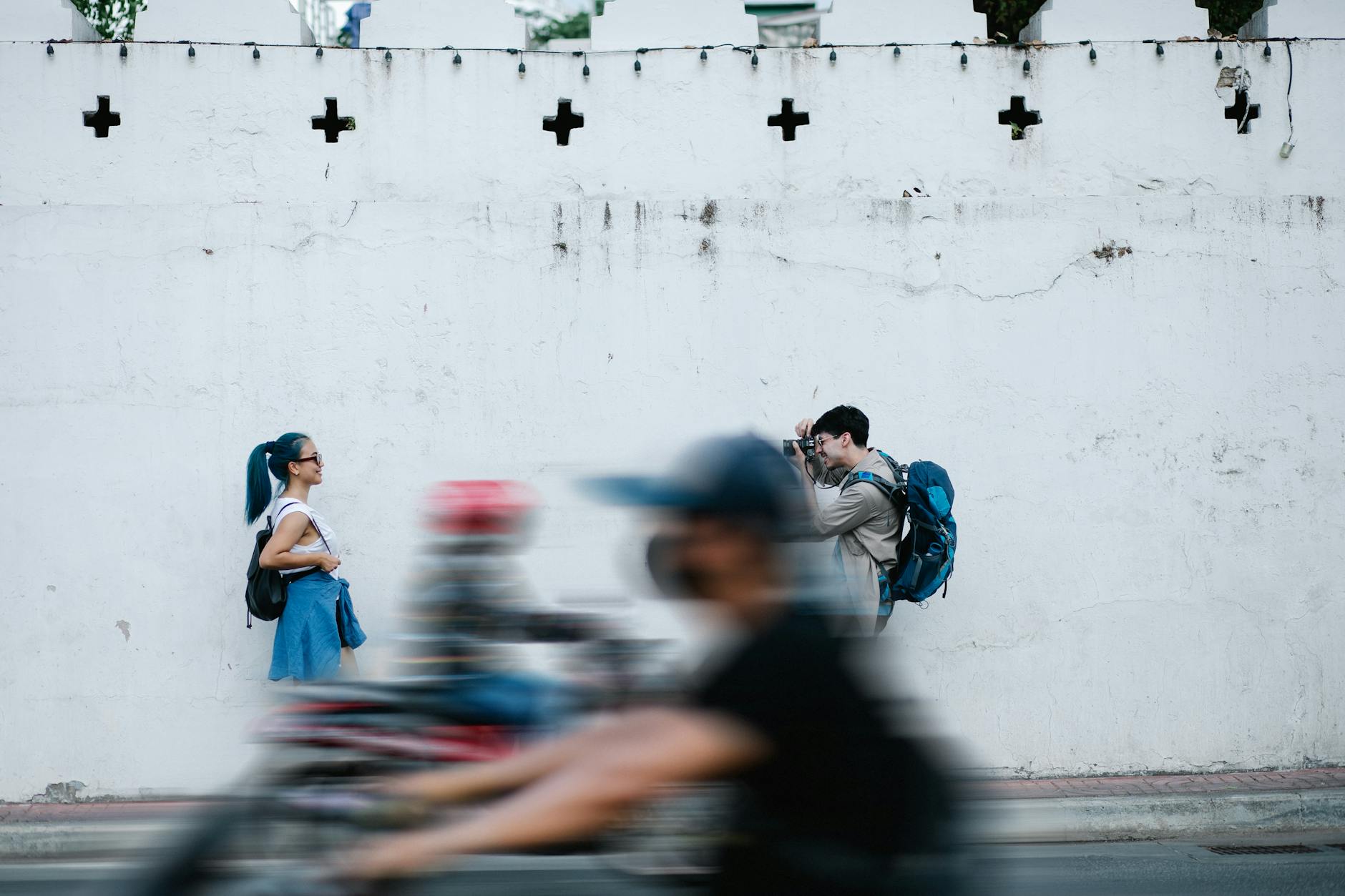 A photographer taking a picture of a woman against a white wall with a passerby in the foreground.