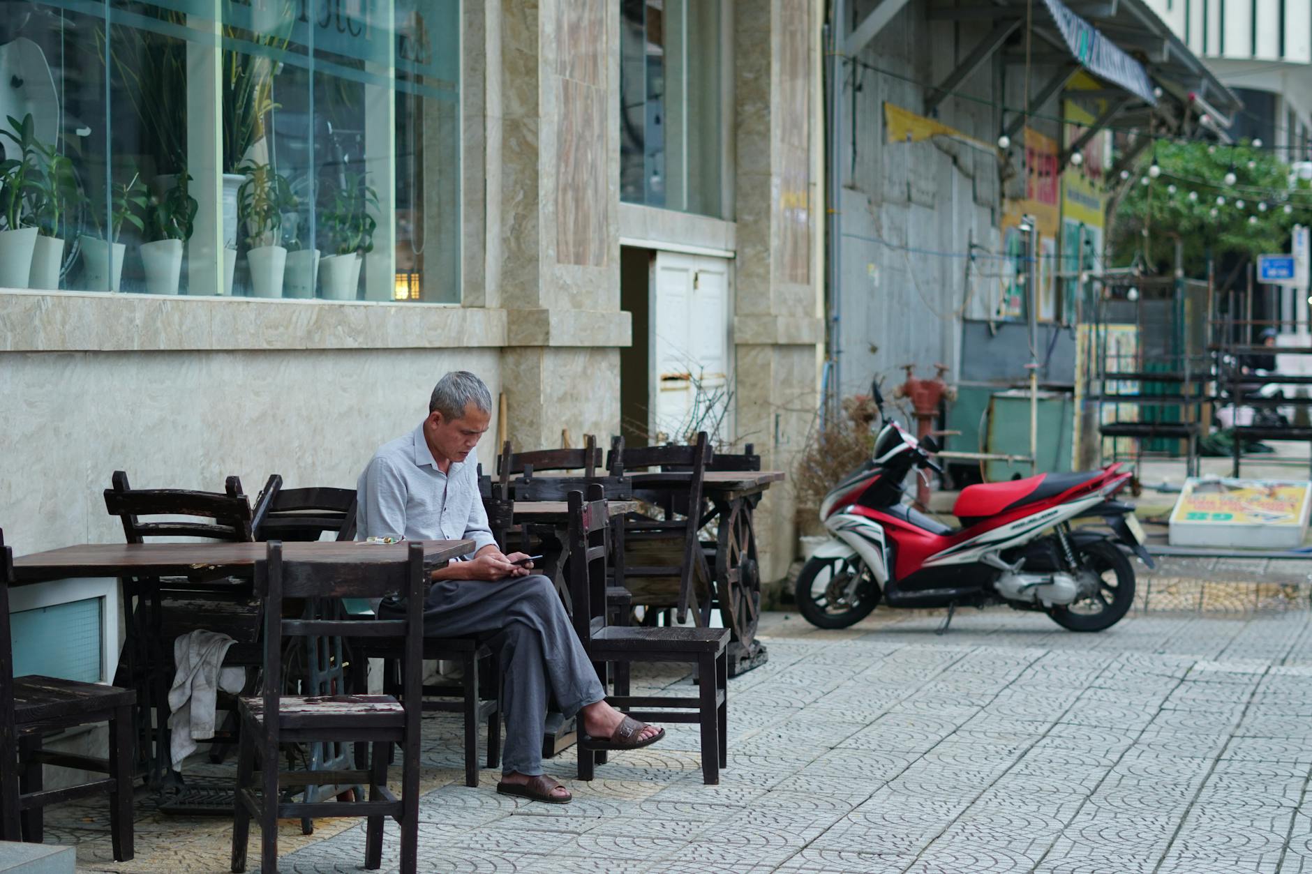 An older man sits outside a cafe using his phone, with wooden chairs and a parked motorcycle nearby.
