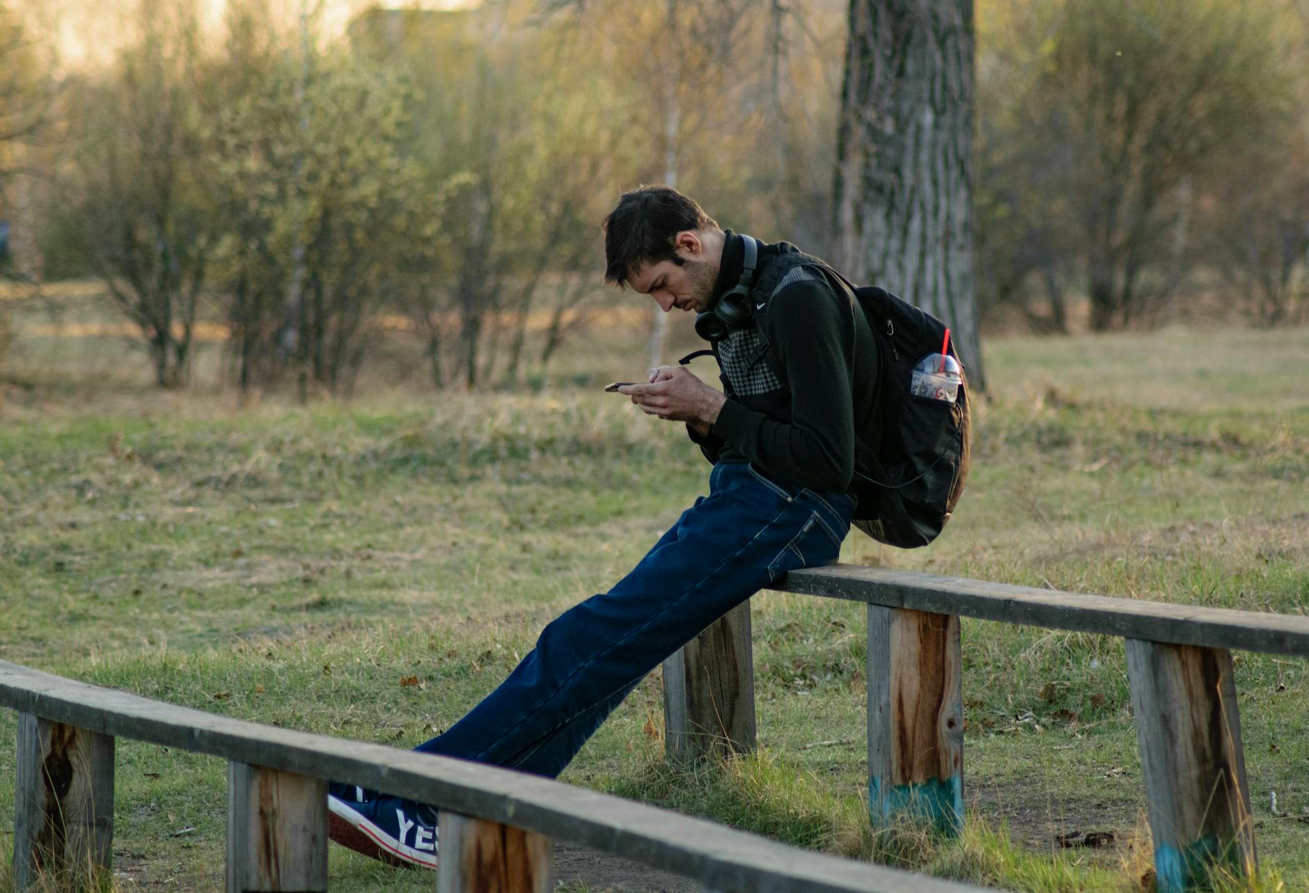 A young man sitting on a bench in a park using his smartphone, wearing headphones.