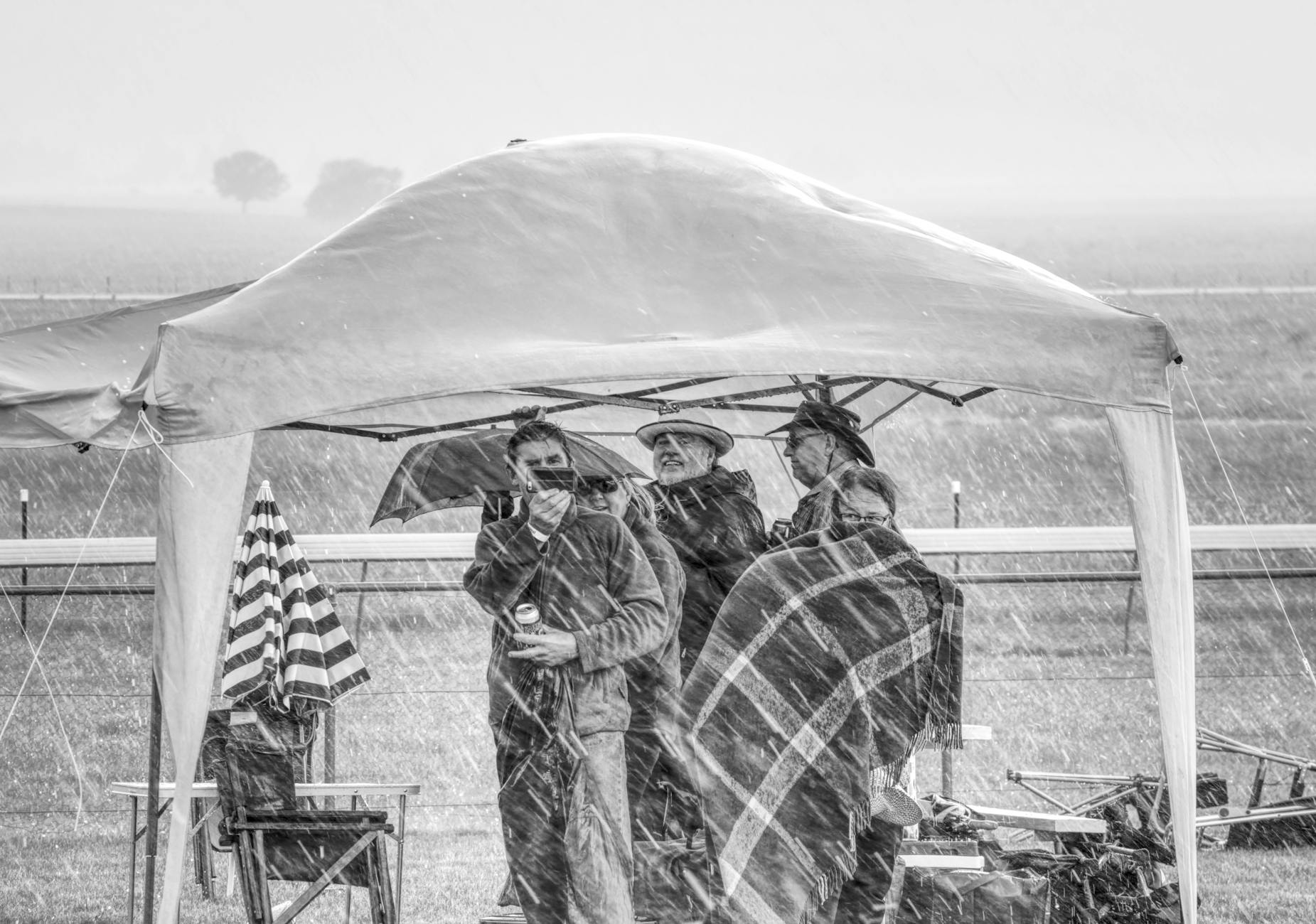 Group of people in rain, huddling under a tent with umbrellas. Black and white photo.