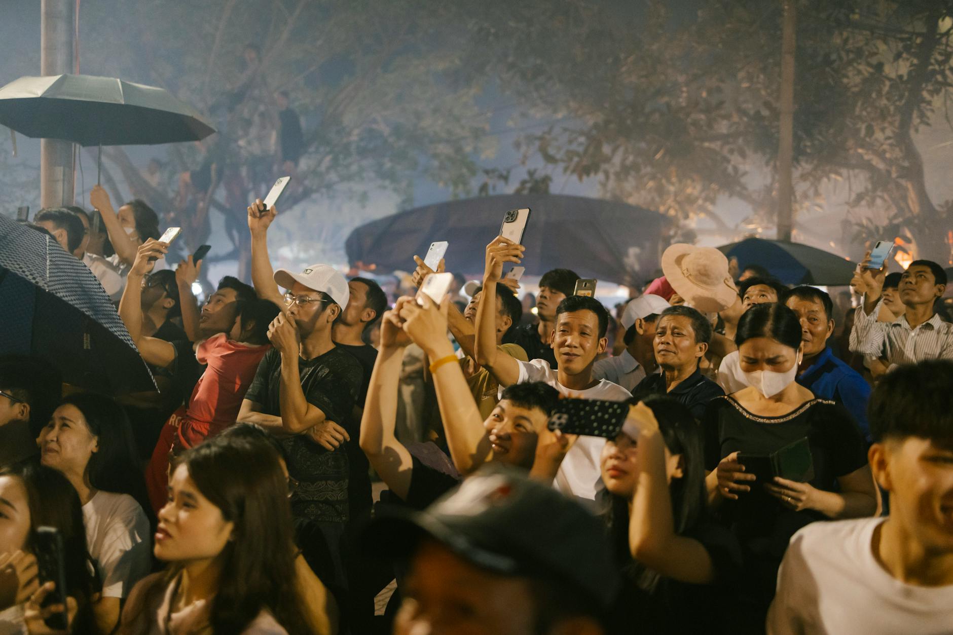 A diverse crowd captures a nighttime event using smartphones and umbrellas, creating a lively atmosphere.