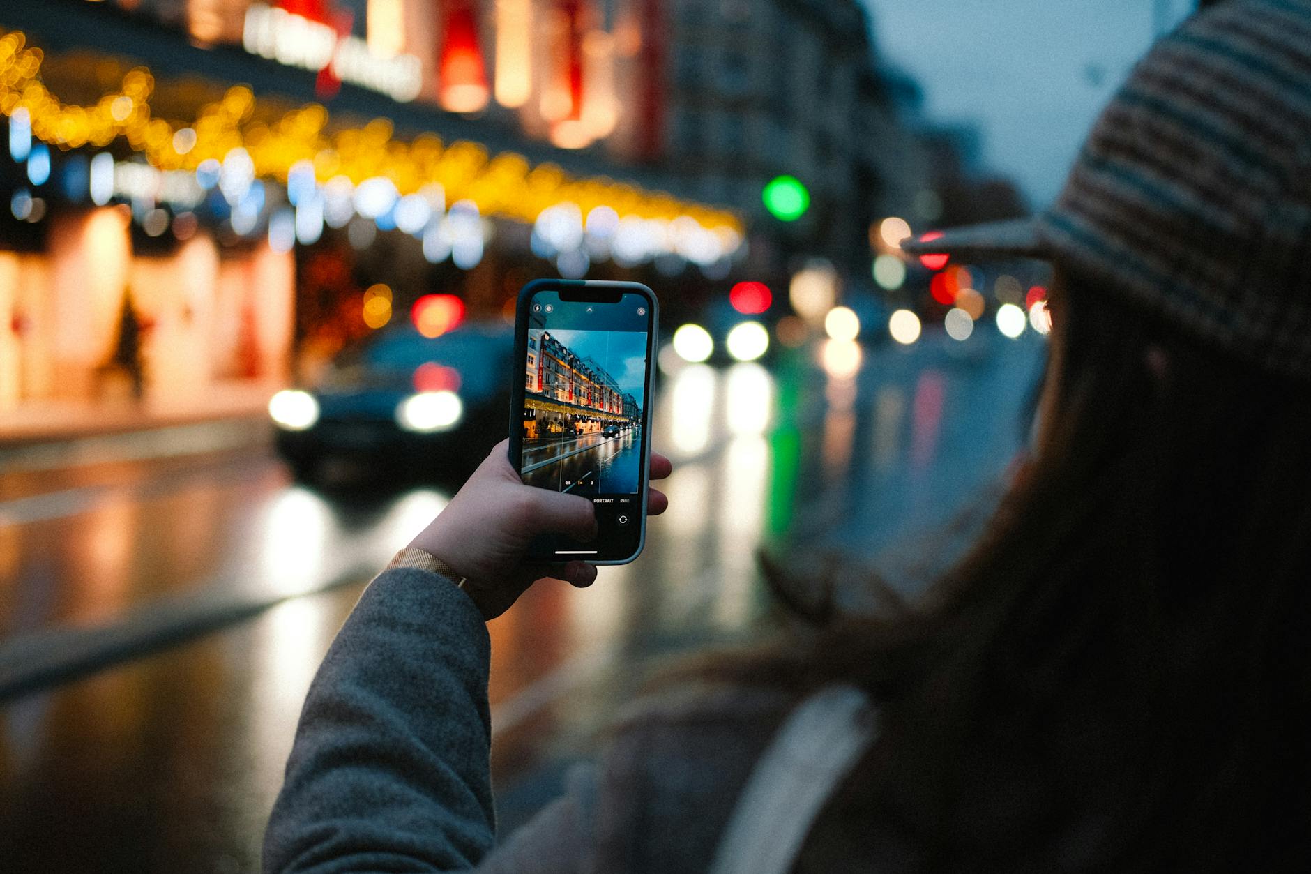 A woman photographs a vibrant city street with glowing lights and passing cars using her smartphone.