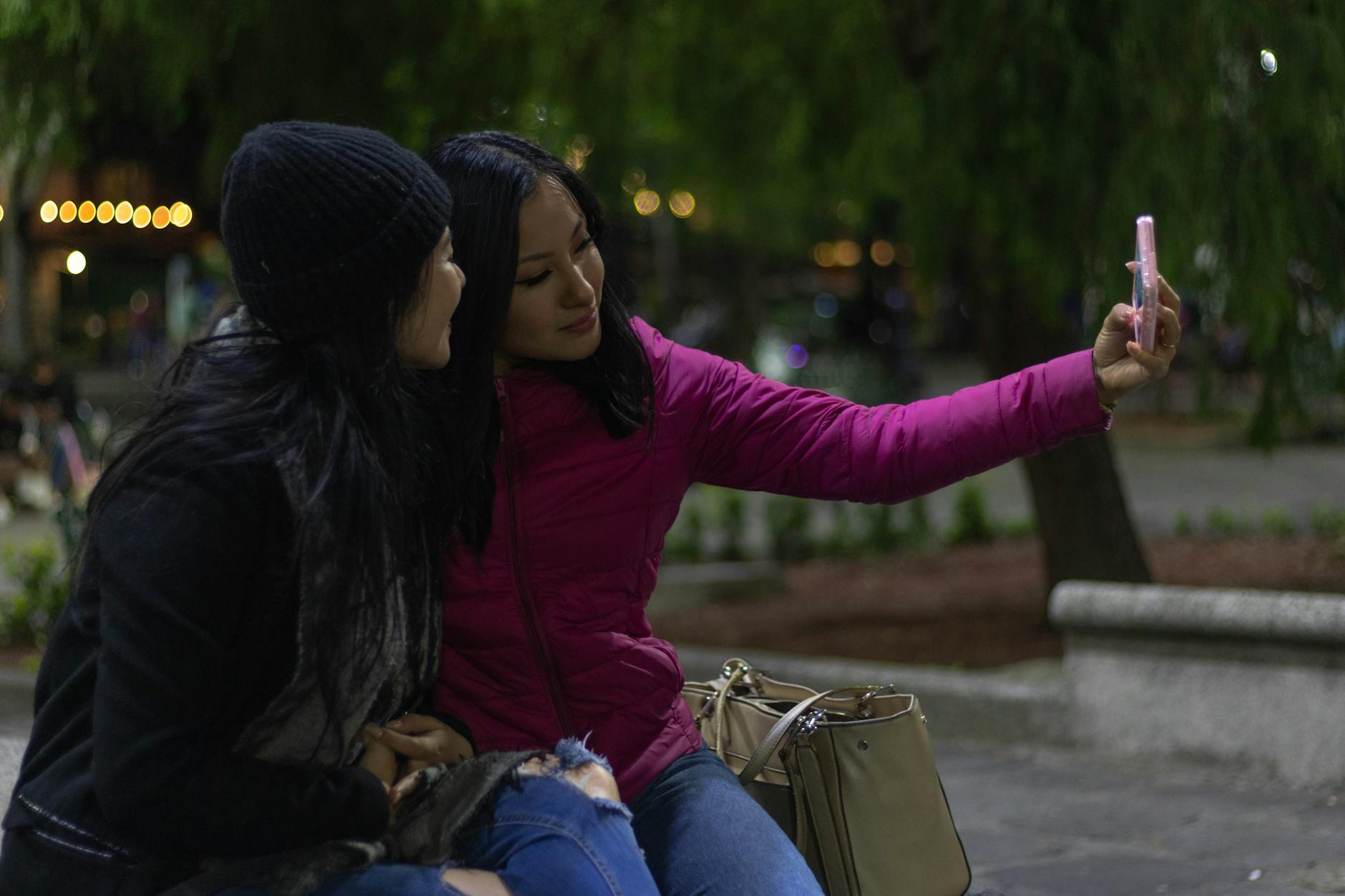 Two women sitting outdoors at night, taking a selfie with a smartphone in a park.