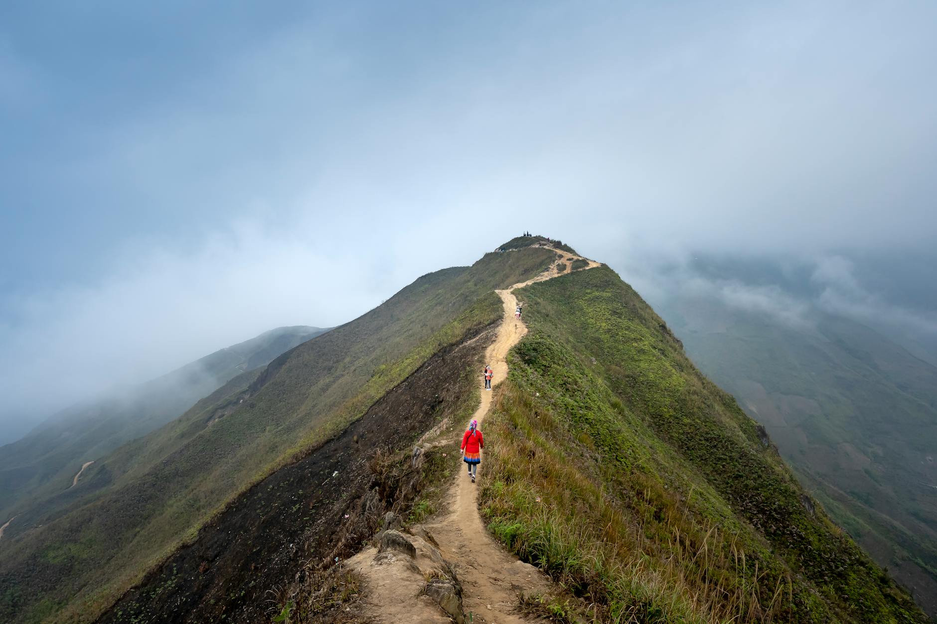 Distant travelers strolling on narrow path on grassy hilltop in highland are under clouds in misty day