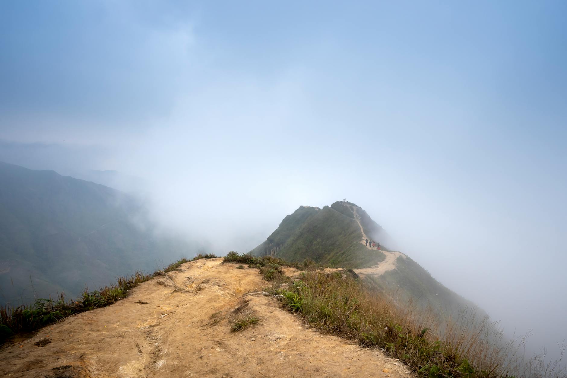 Distant tourists walking on steep ridge in mountainous valley covered with clouds in haze