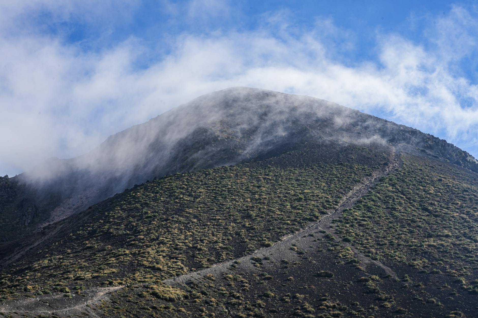A remote mountain peak with a cloud veil and rugged trail under a bright sky.