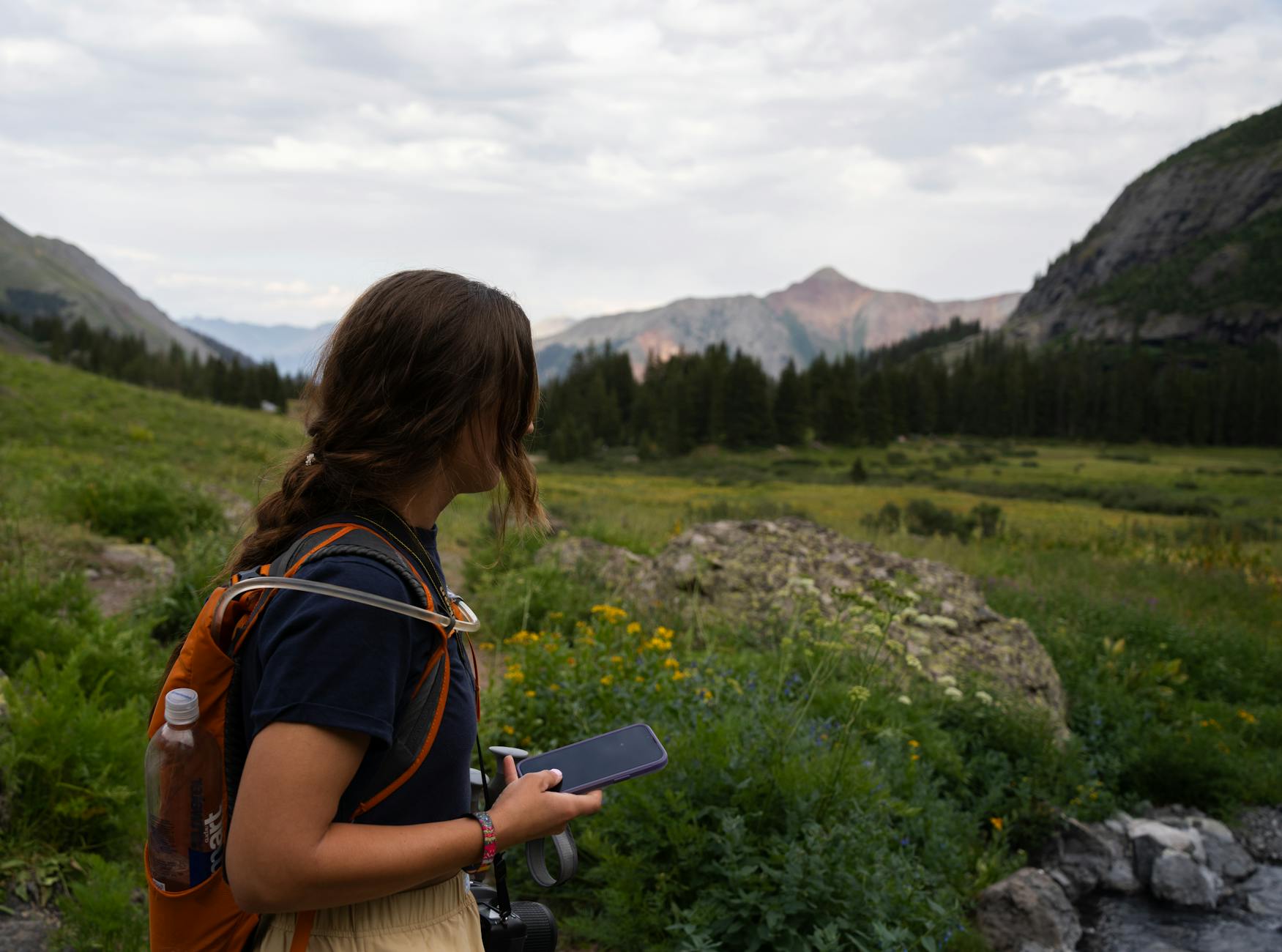 A young woman hikes through the green landscape of Colorado, exploring nature with her camera and phone.