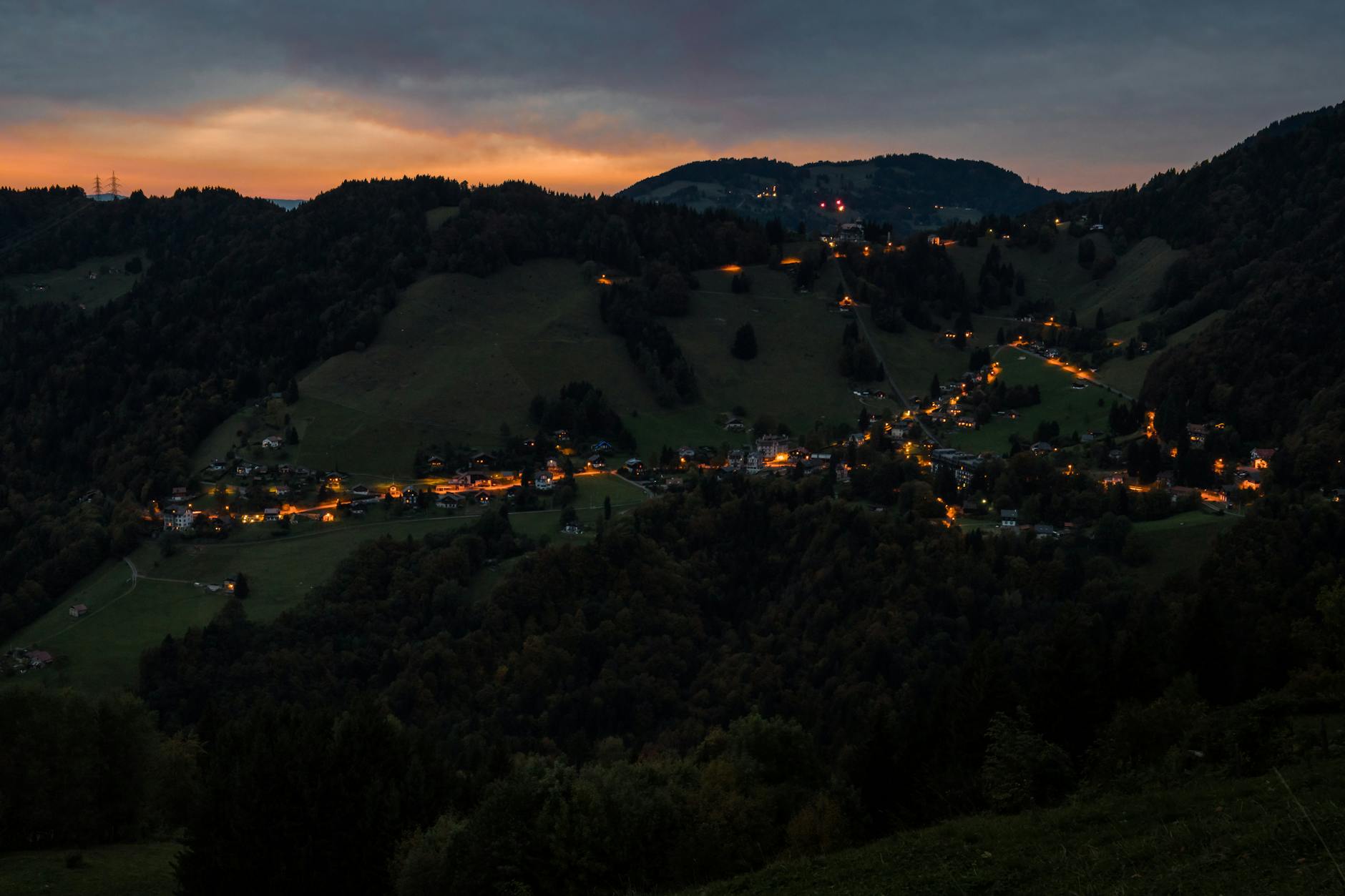 Elevated view of a village nestled in dark hills at twilight, lights glowing, under a cloudy sky.