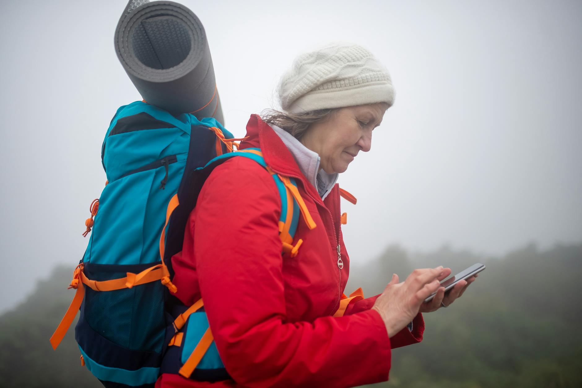 Elderly woman in red jacket checks phone while hiking in foggy scenery outdoors.