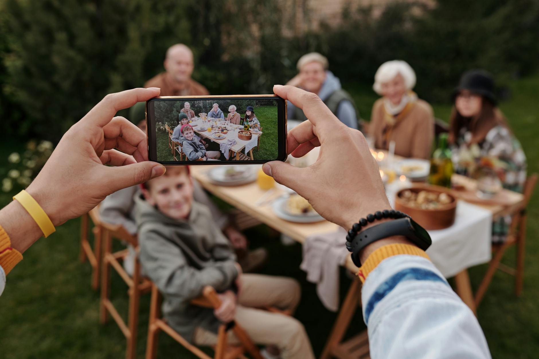 Faceless person with fitness bracelet taking photo with smartphone of big multigenerational family dinner served outside