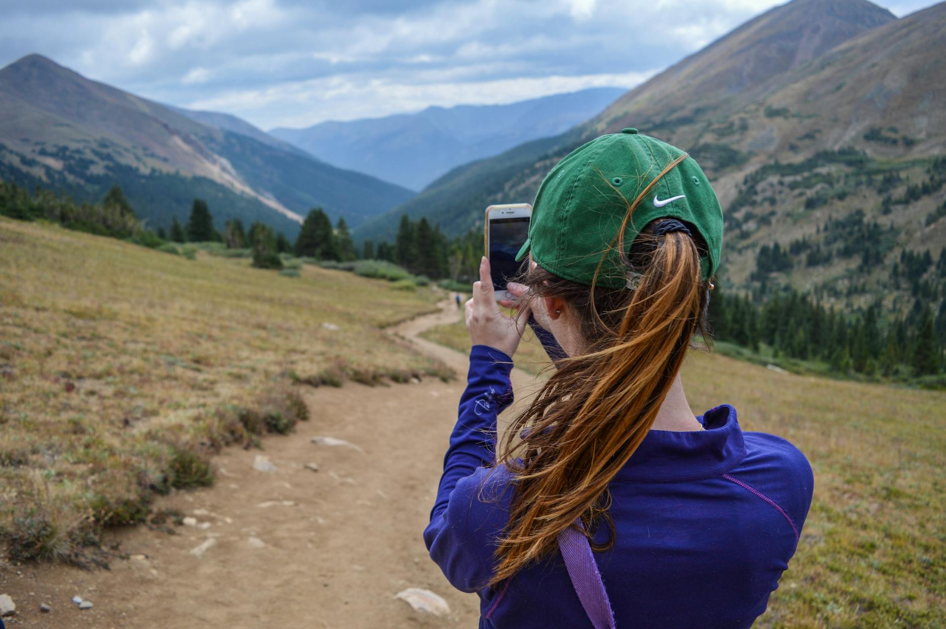 Woman capturing scenic mountain view with smartphone while hiking on a trail.