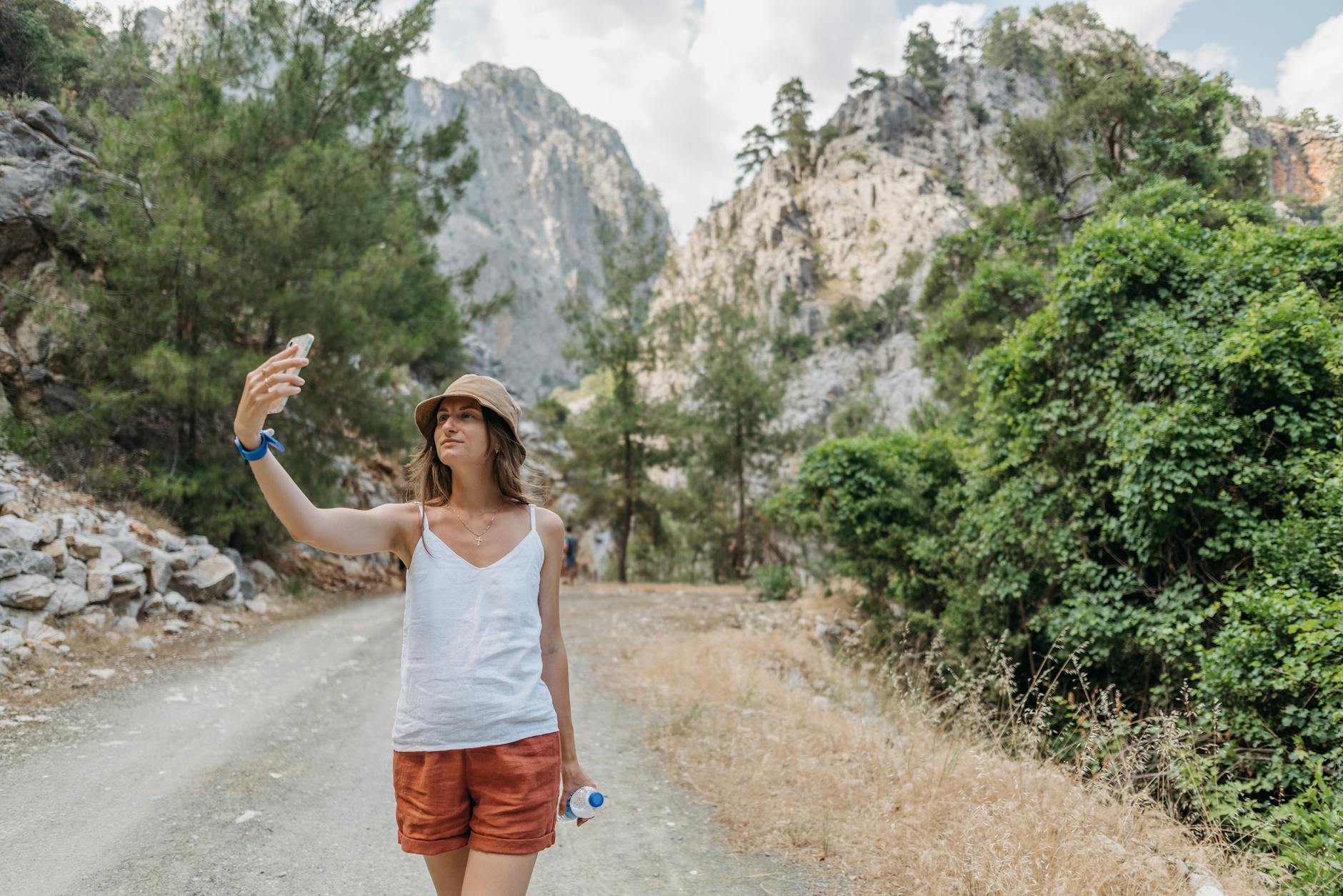 Woman taking a selfie on a scenic mountain trail surrounded by lush greenery and rocky cliffs.