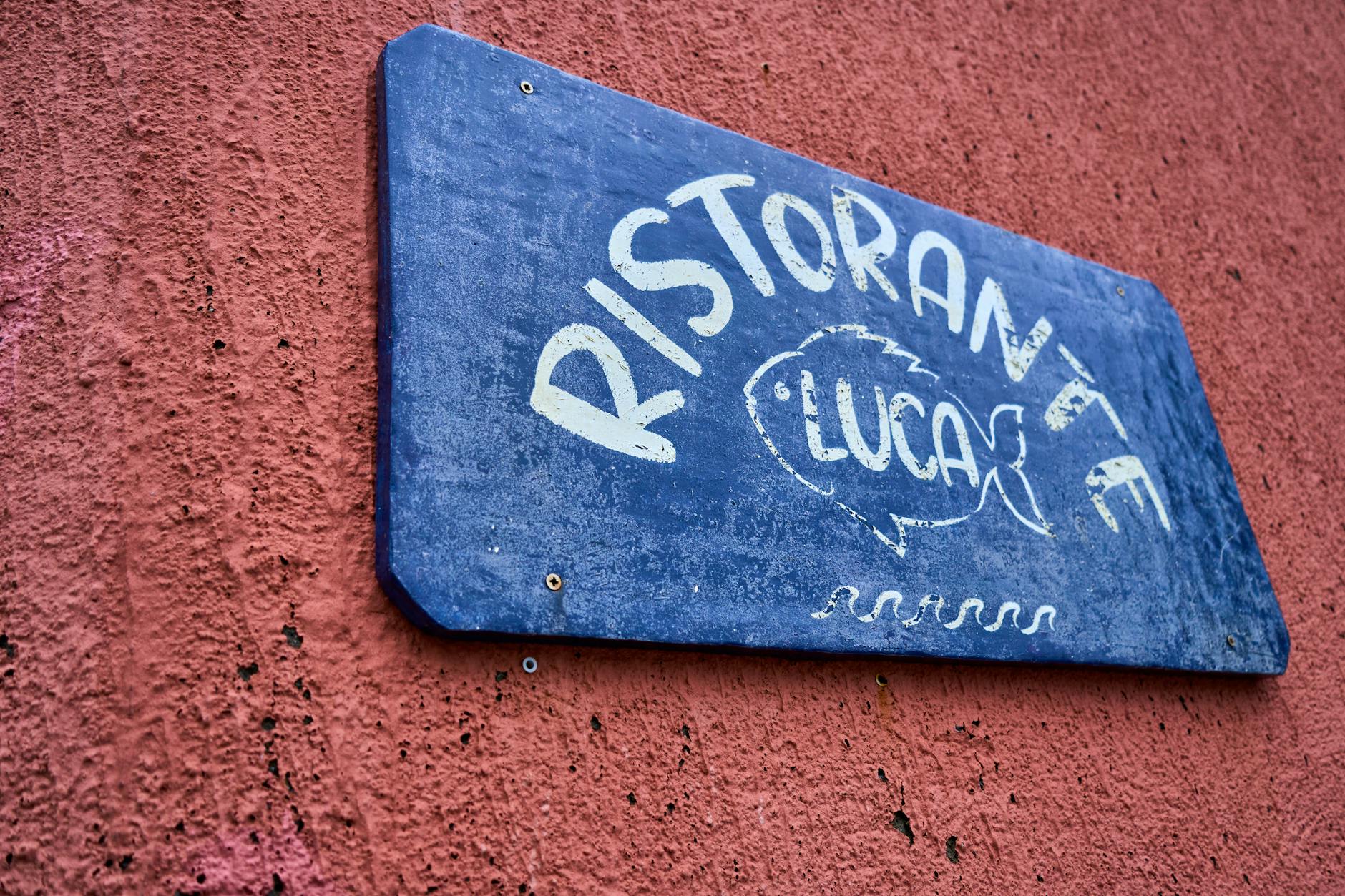 Blue restaurant sign 'RISTORANTE LUCA' on a red stucco wall, with fish illustration.