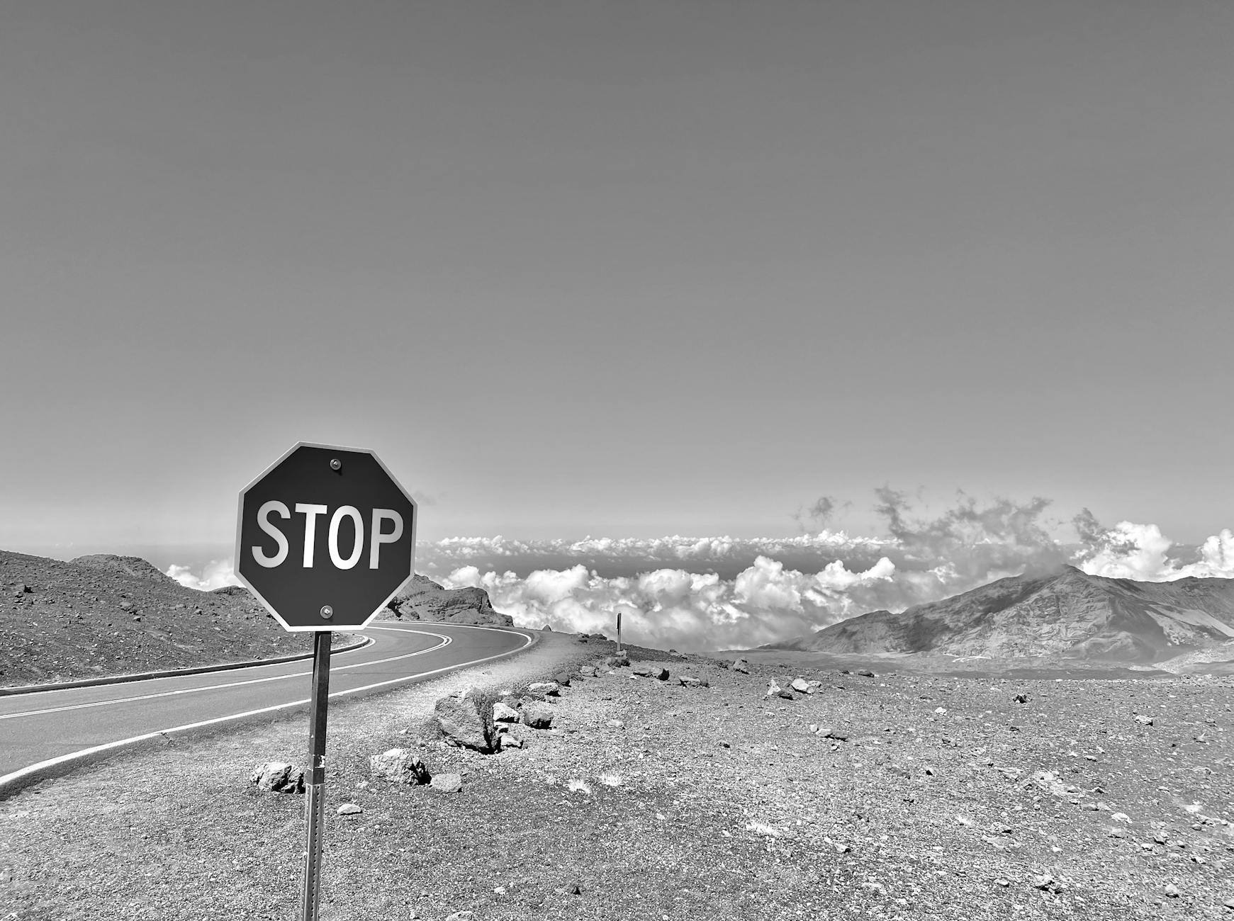 Black and white photo of a stop sign on a mountain road with clouds in the distance.