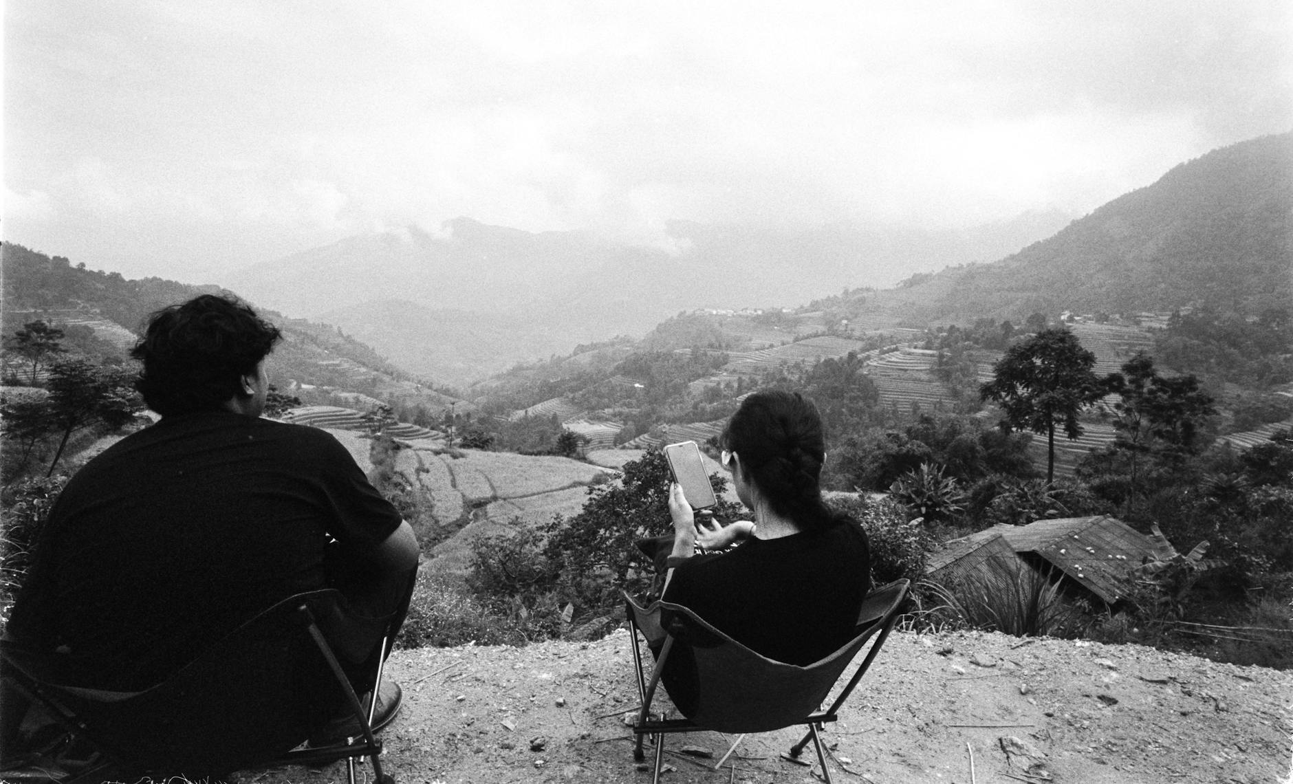 Black and white photo of couple using phones on a hillside, overlooking rural landscape.