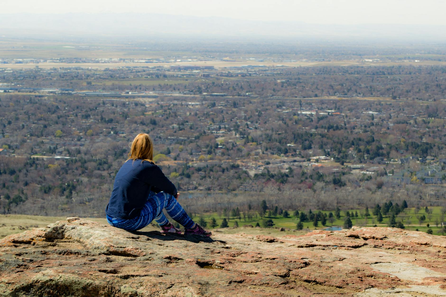 A woman sits on a rocky hill with a vast view of the city and nature below, enjoying a peaceful moment.