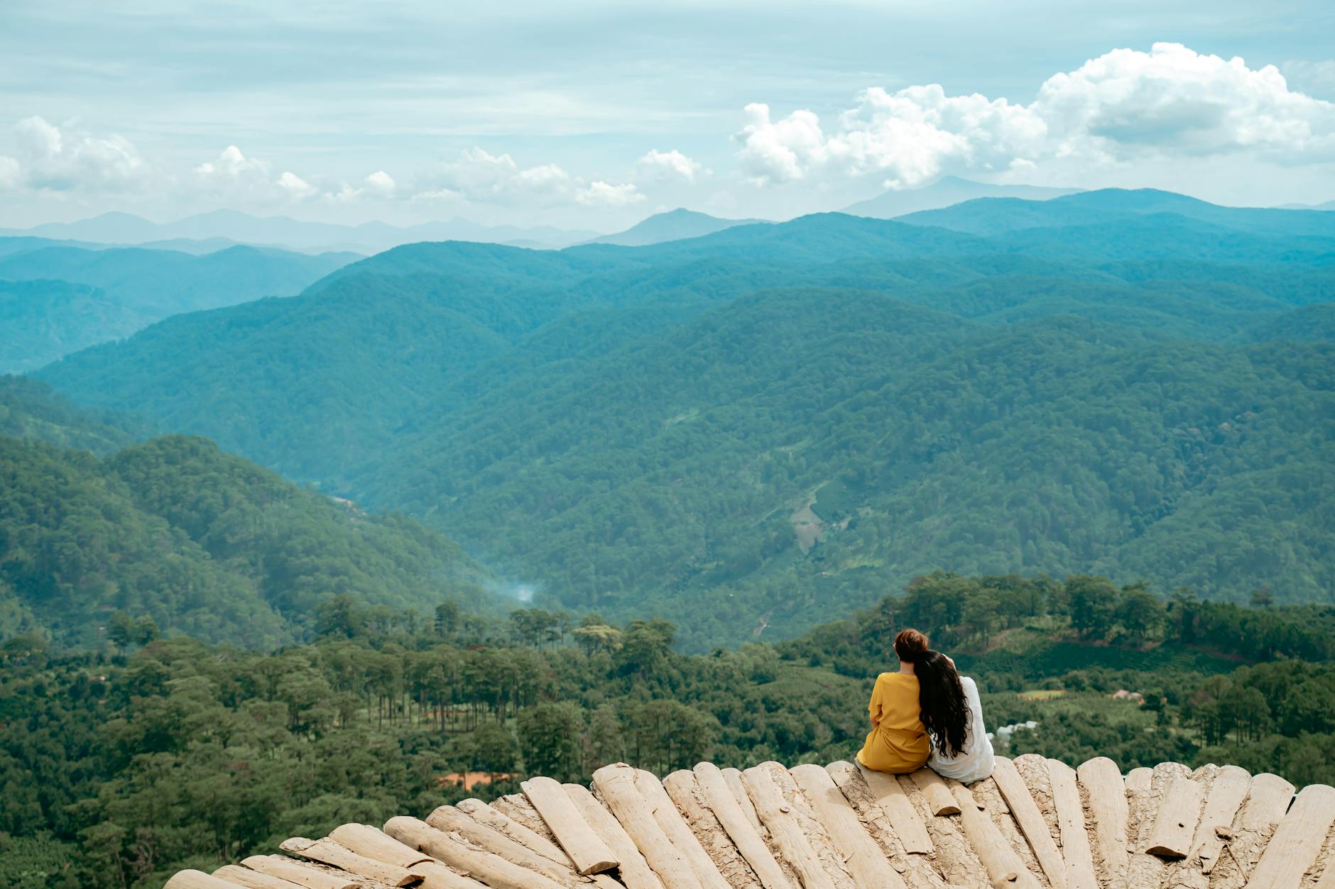 Back view of anonymous tourists cuddling while sitting on viewpoint and admiring picturesque mountain valley covered with lush green vegetation