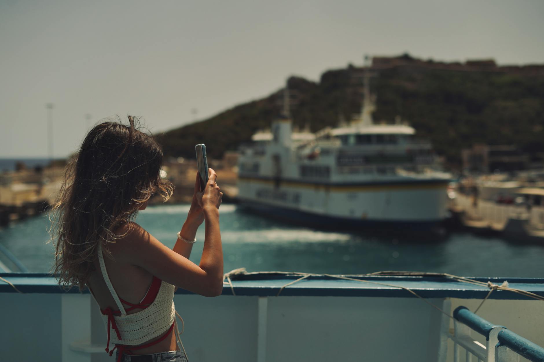 A young woman uses her smartphone to capture a ferry scene on a sunny day.