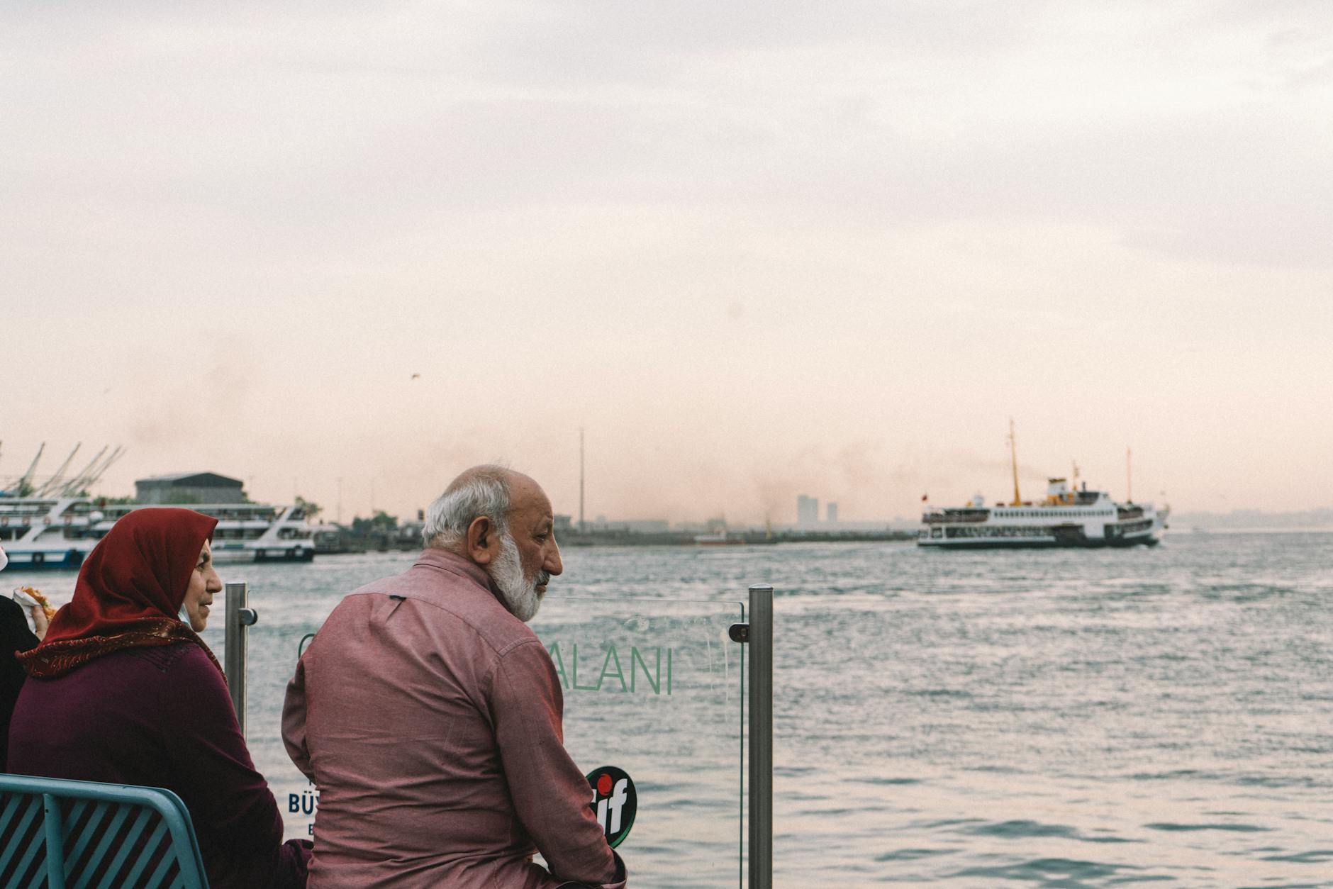 An elderly couple sitting by the seaside watching a ferry under cloudy skies.