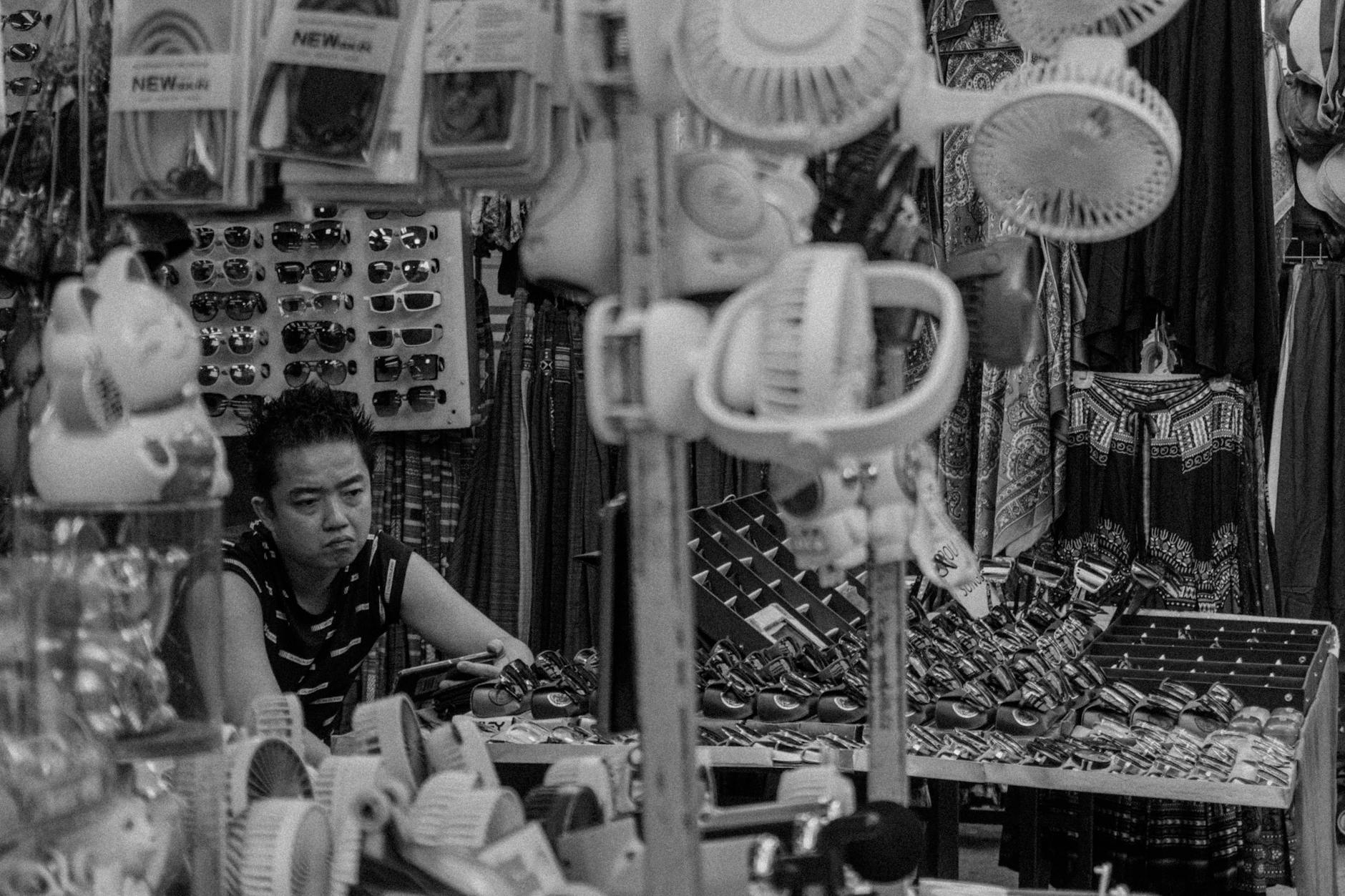 Black and white photo of a busy street market with a vendor surrounded by various goods.