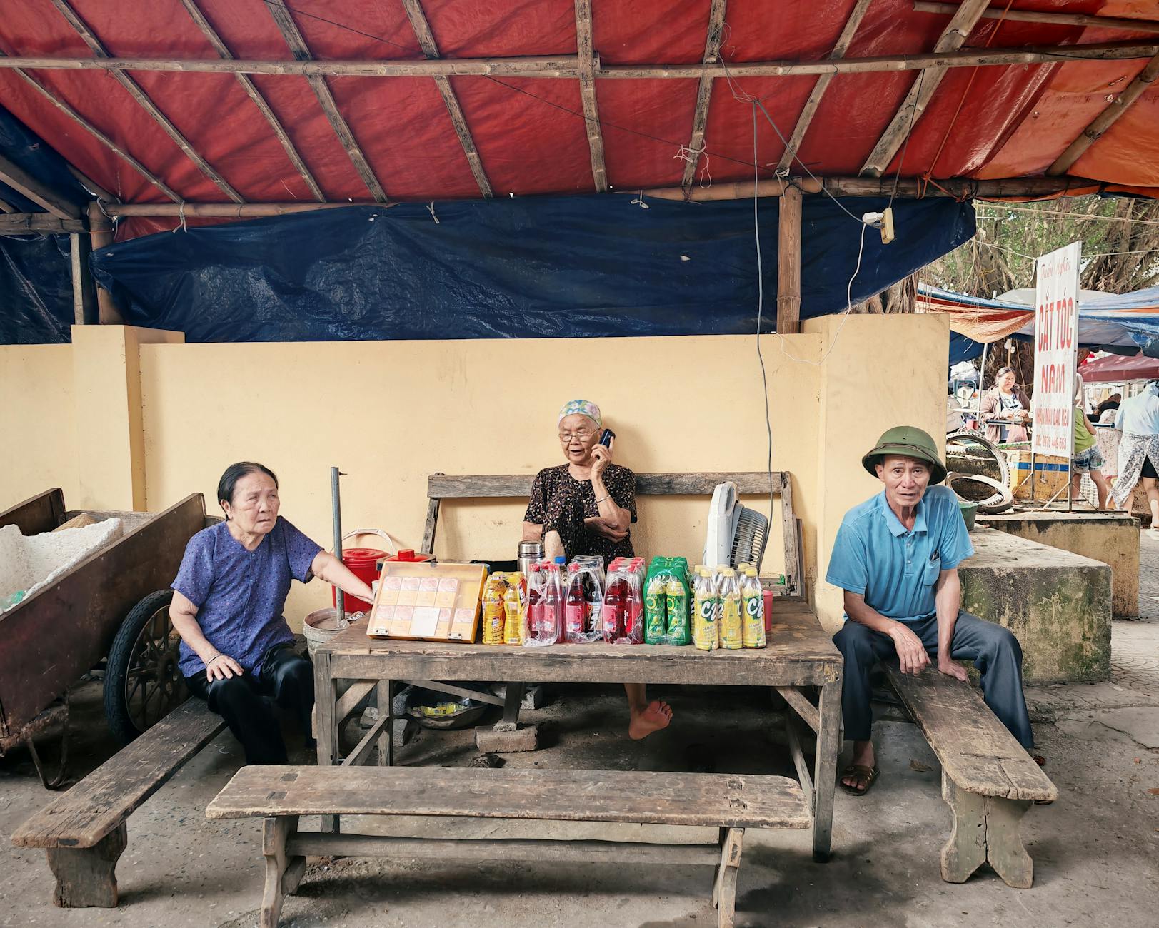 Three elderly vendors sit at a street market stall displaying bottled drinks.
