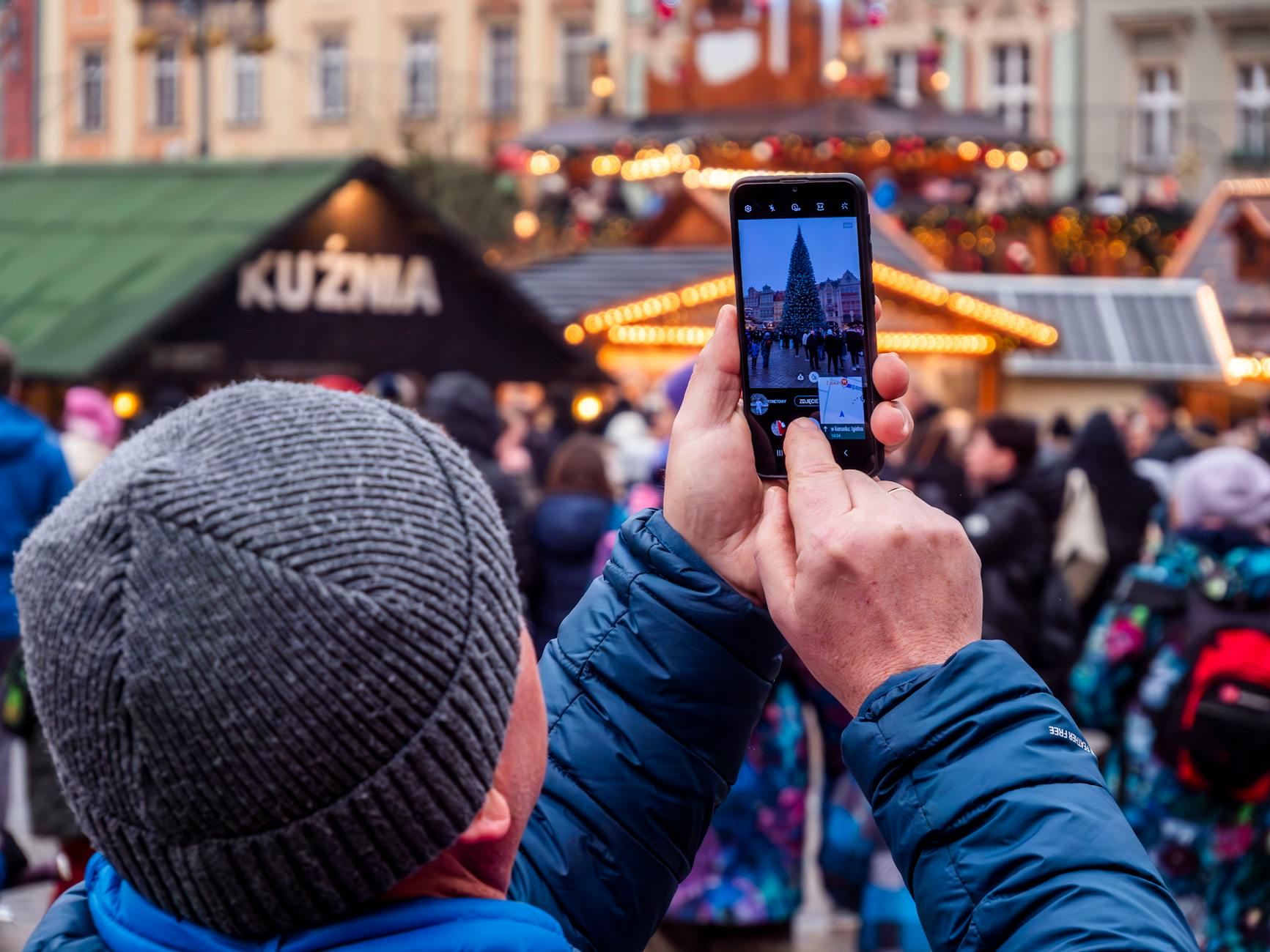 Man photographing Christmas market decorations with phone in winter crowd.