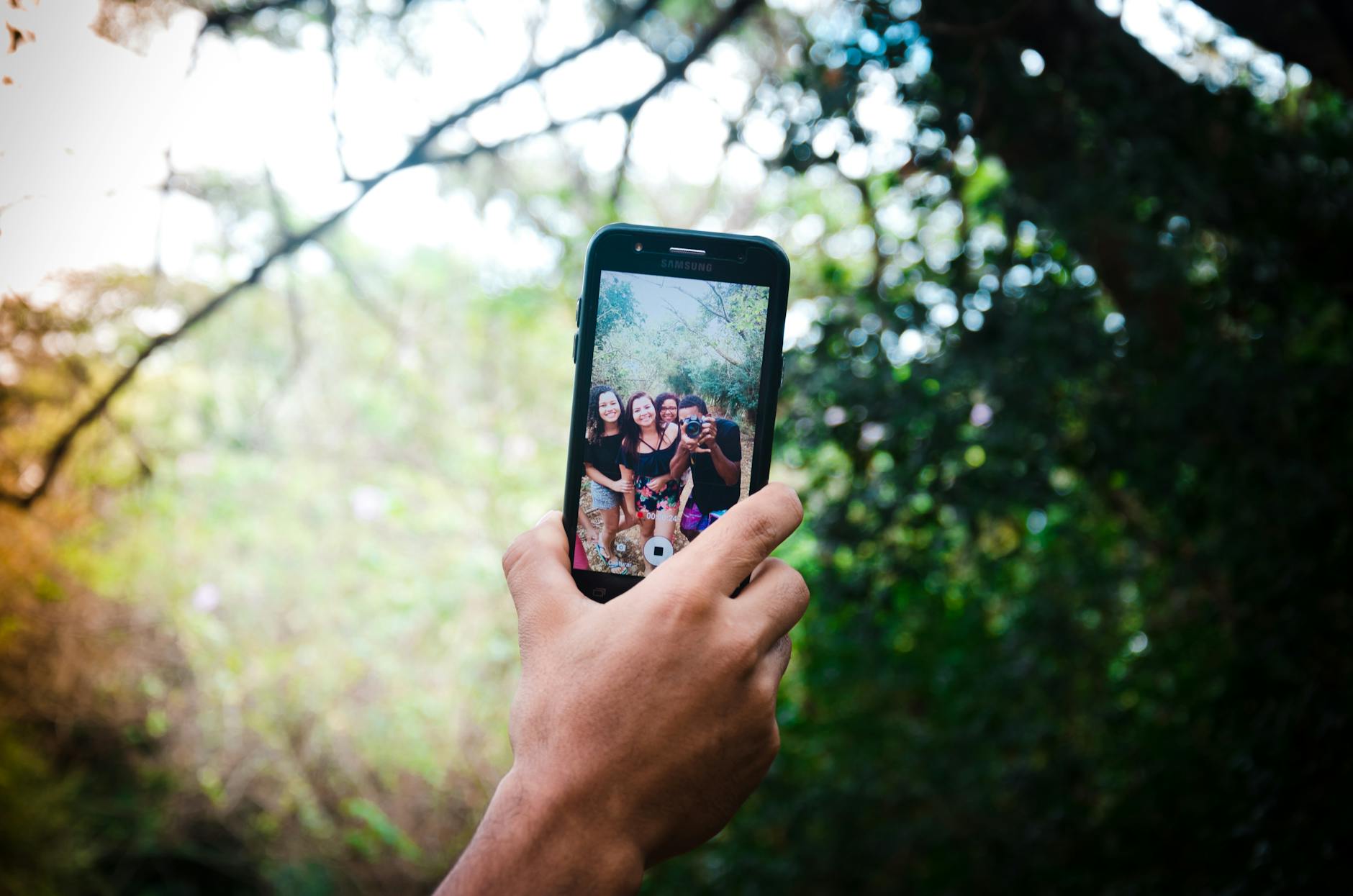 A hand holds a smartphone capturing a selfie of friends in a lush outdoor setting.