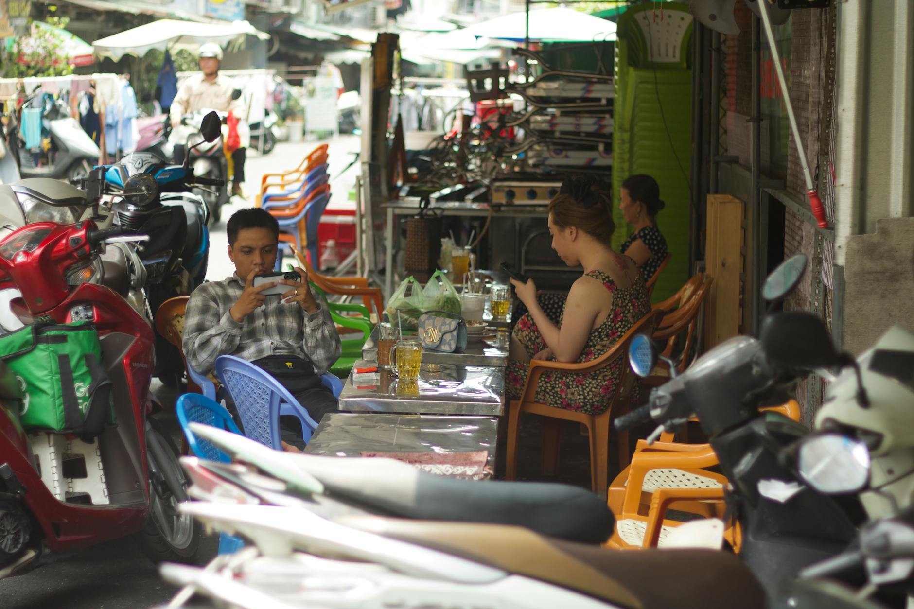 A lively street scene featuring people dining and using phones at an outdoor cafe in Hồ Chí Minh City, Vietnam.