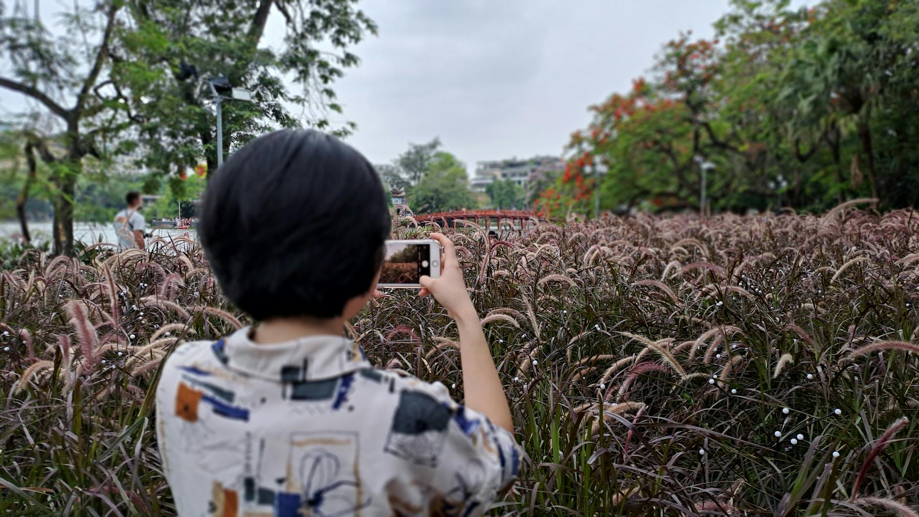 A woman with short black hair photographs lush park scenery using her smartphone.