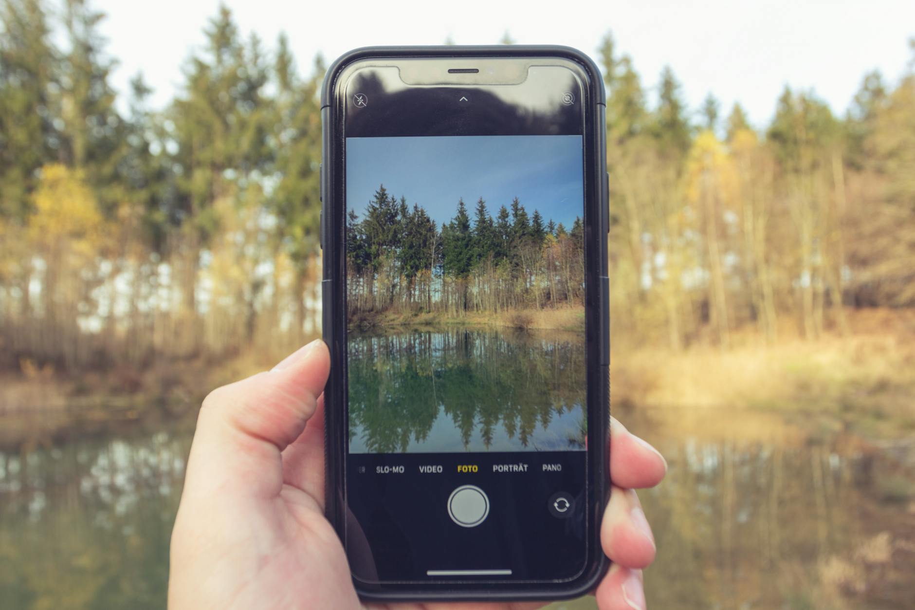 Crop anonymous person taking photo of forest and calm water of river with mobile phone on blurred background of nature
