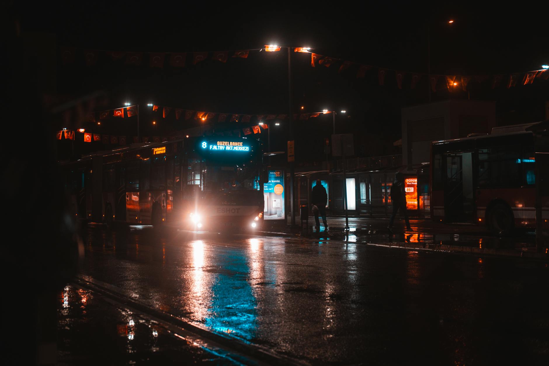 A city bus illuminated in the night at a busy station with wet streets.