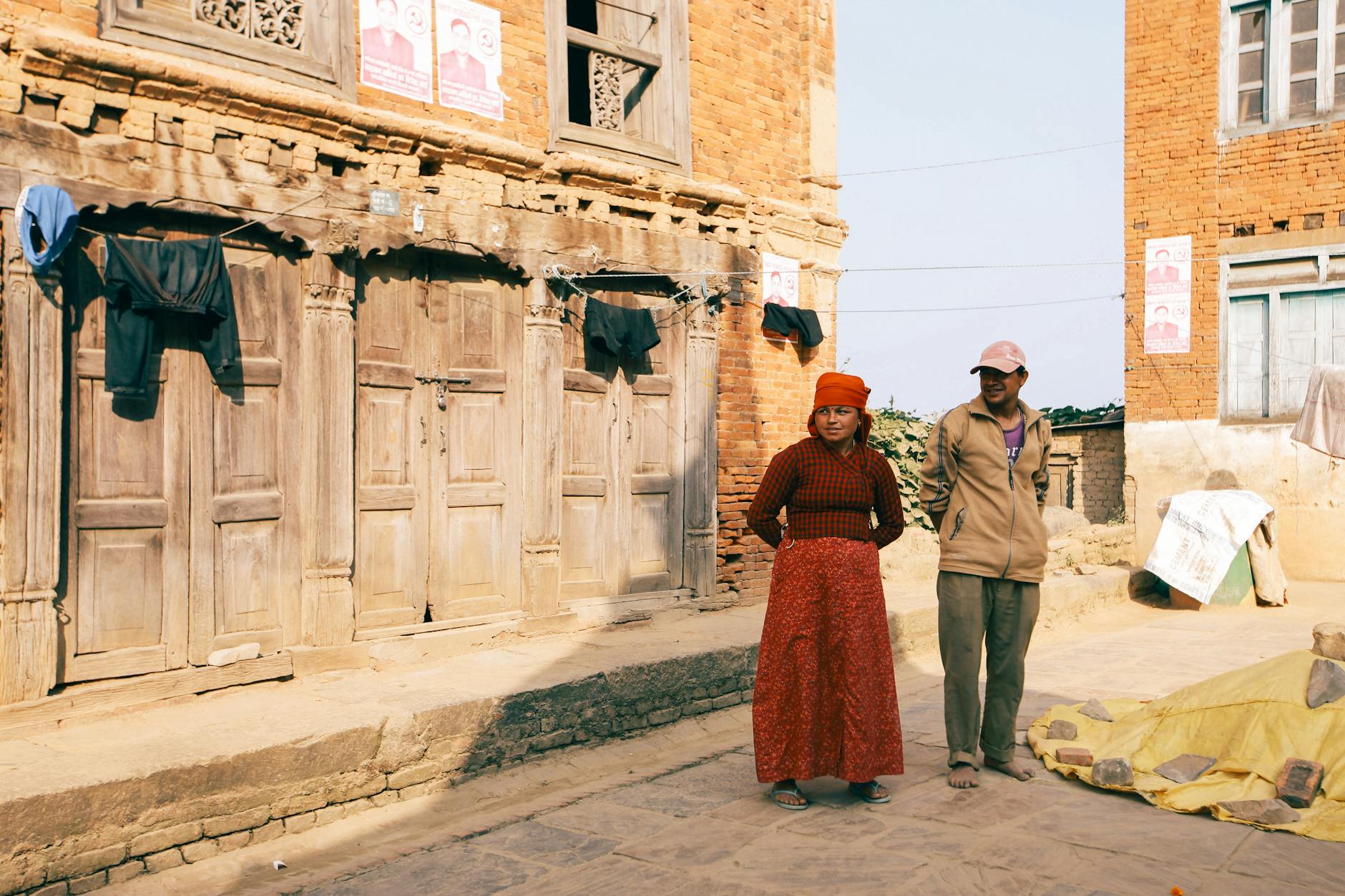 Man and woman in traditional attire stand by rustic red brick homes in a village.