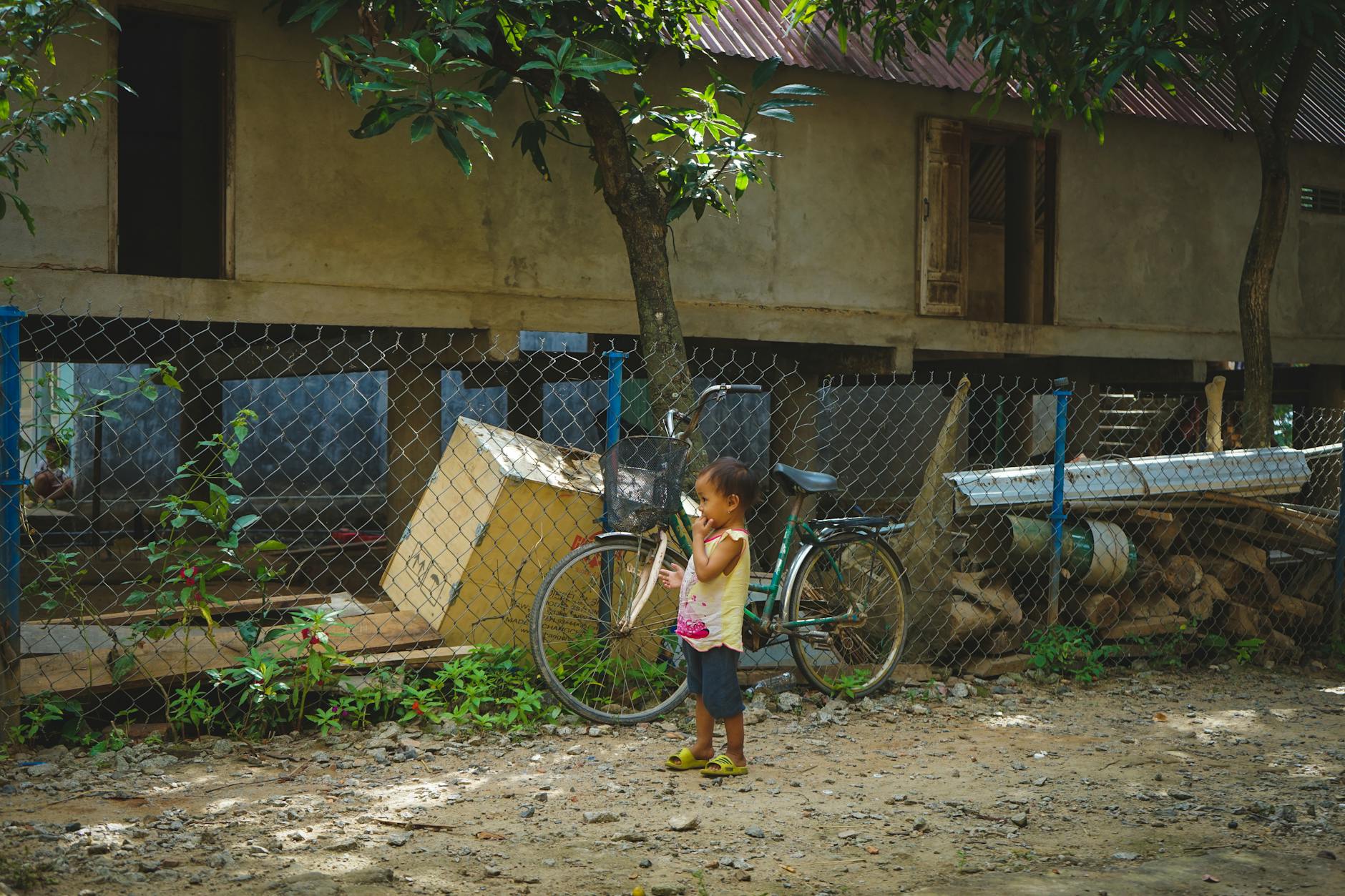 A young child stands near a bicycle in a rural village setting, surrounded by trees and rustic buildings.