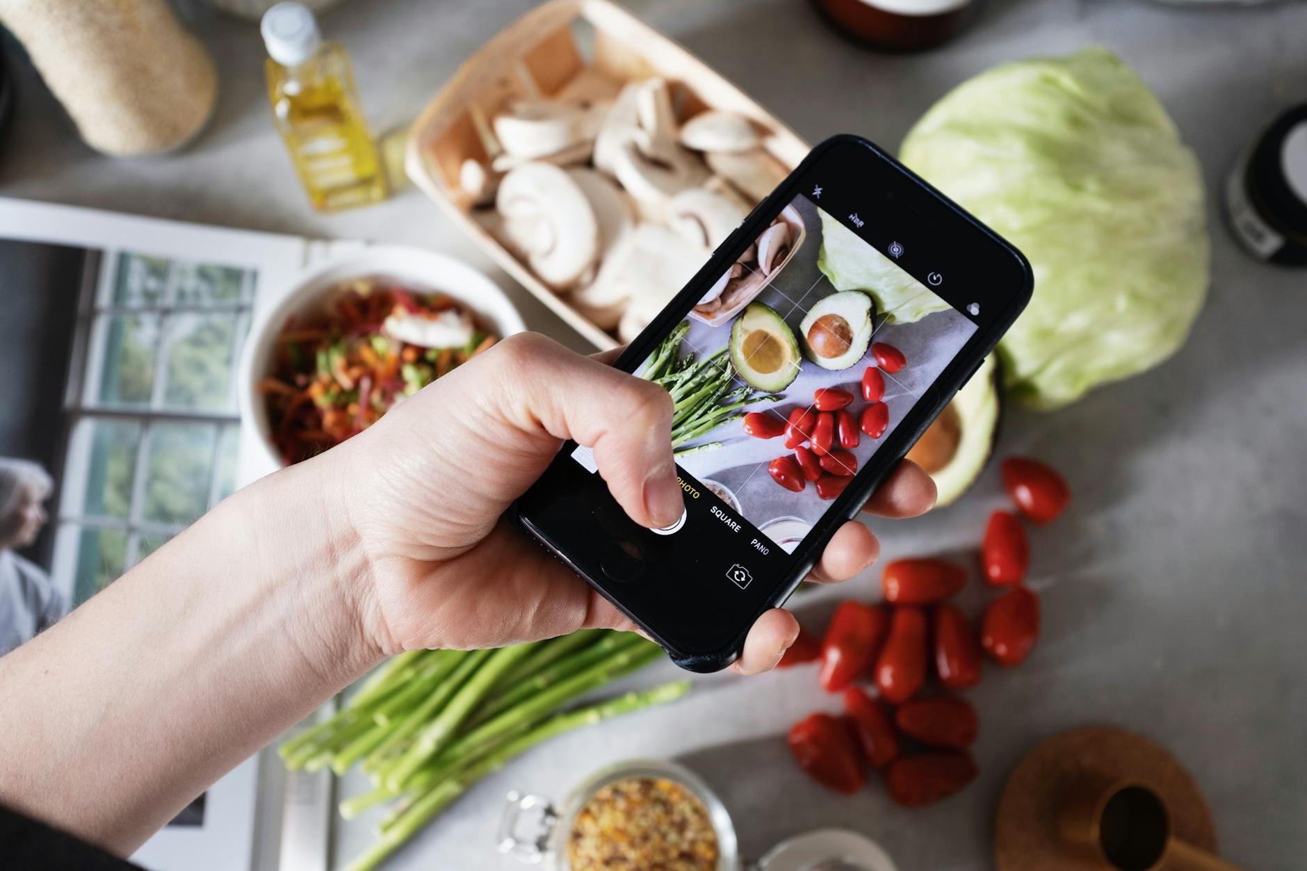 Hand holding smartphone capturing photo of fresh vegetables on a kitchen counter.