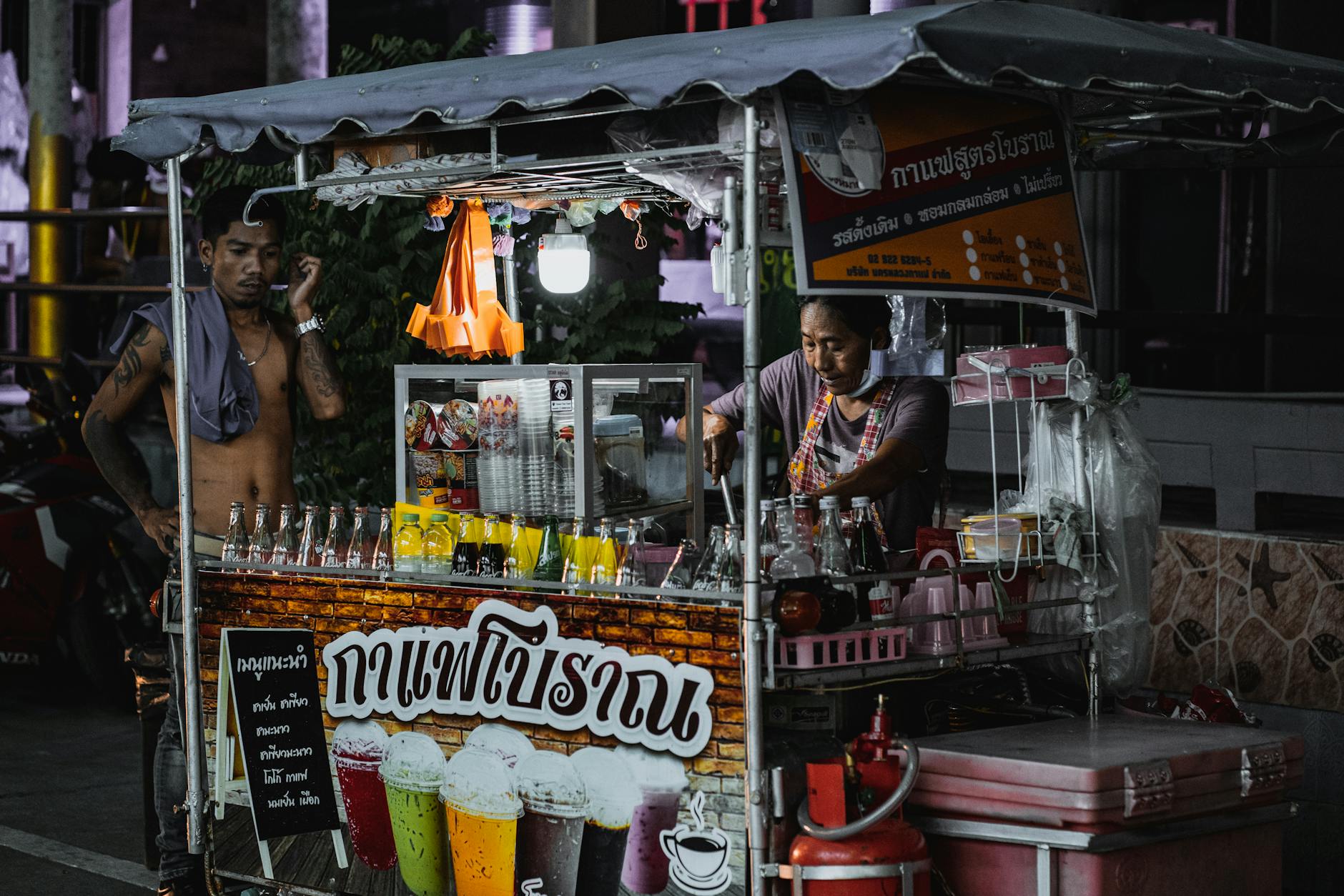 Two vendors operating a colorful beverage cart in Bangkok at night.