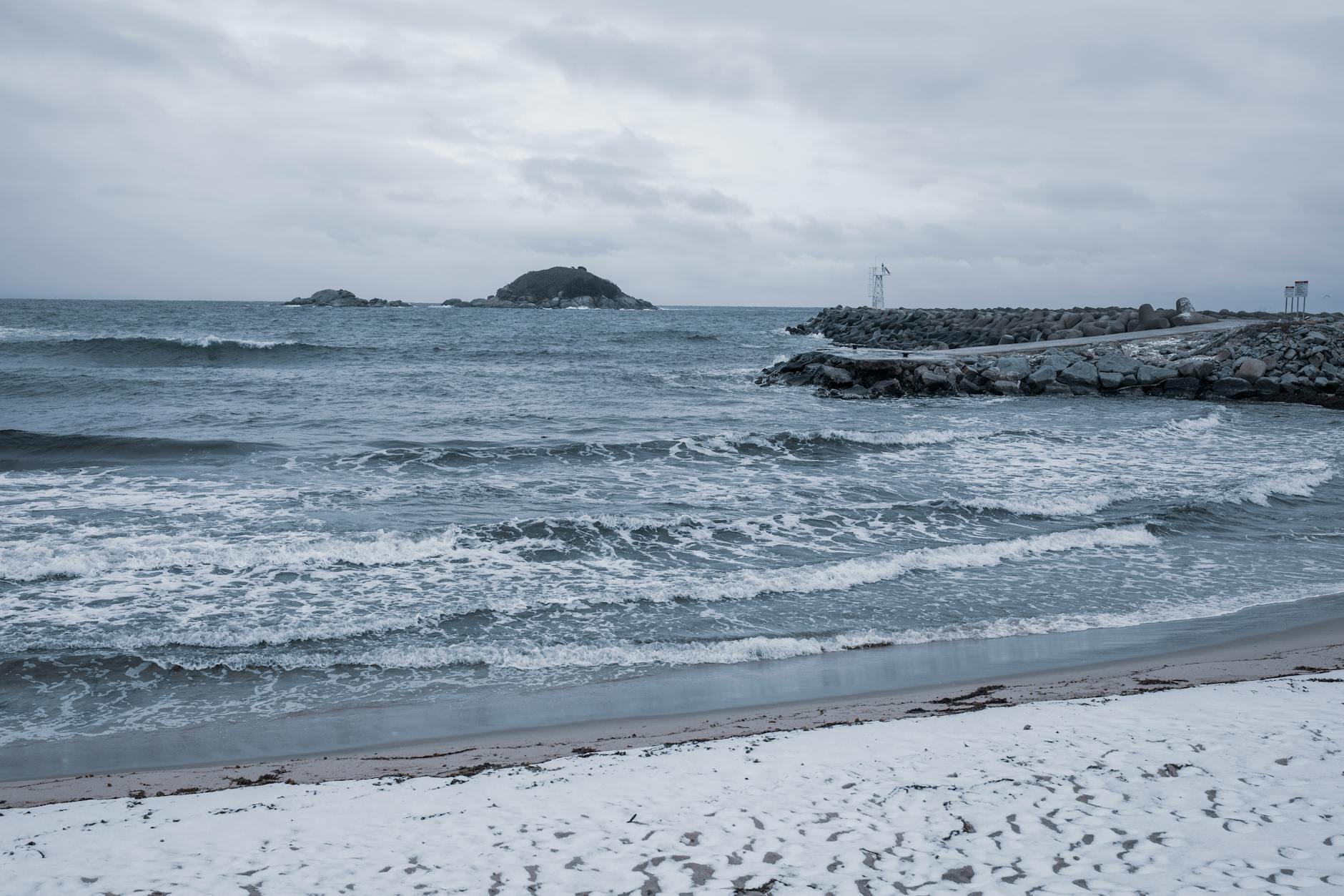 A tranquil snowy beach with waves and a distant island under overcast skies.