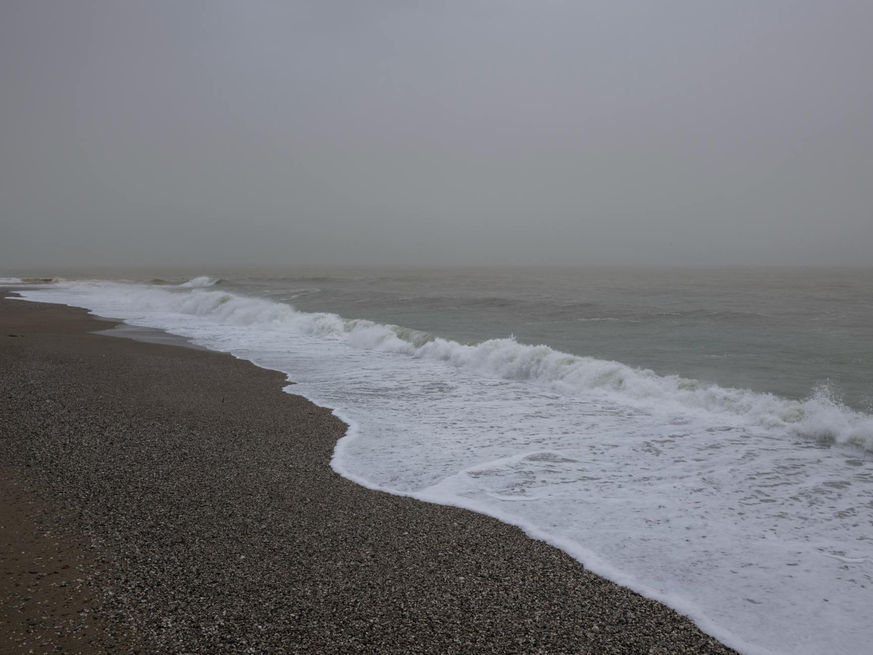 A serene winter seascape with foggy weather and gentle waves on a pebbly shoreline.