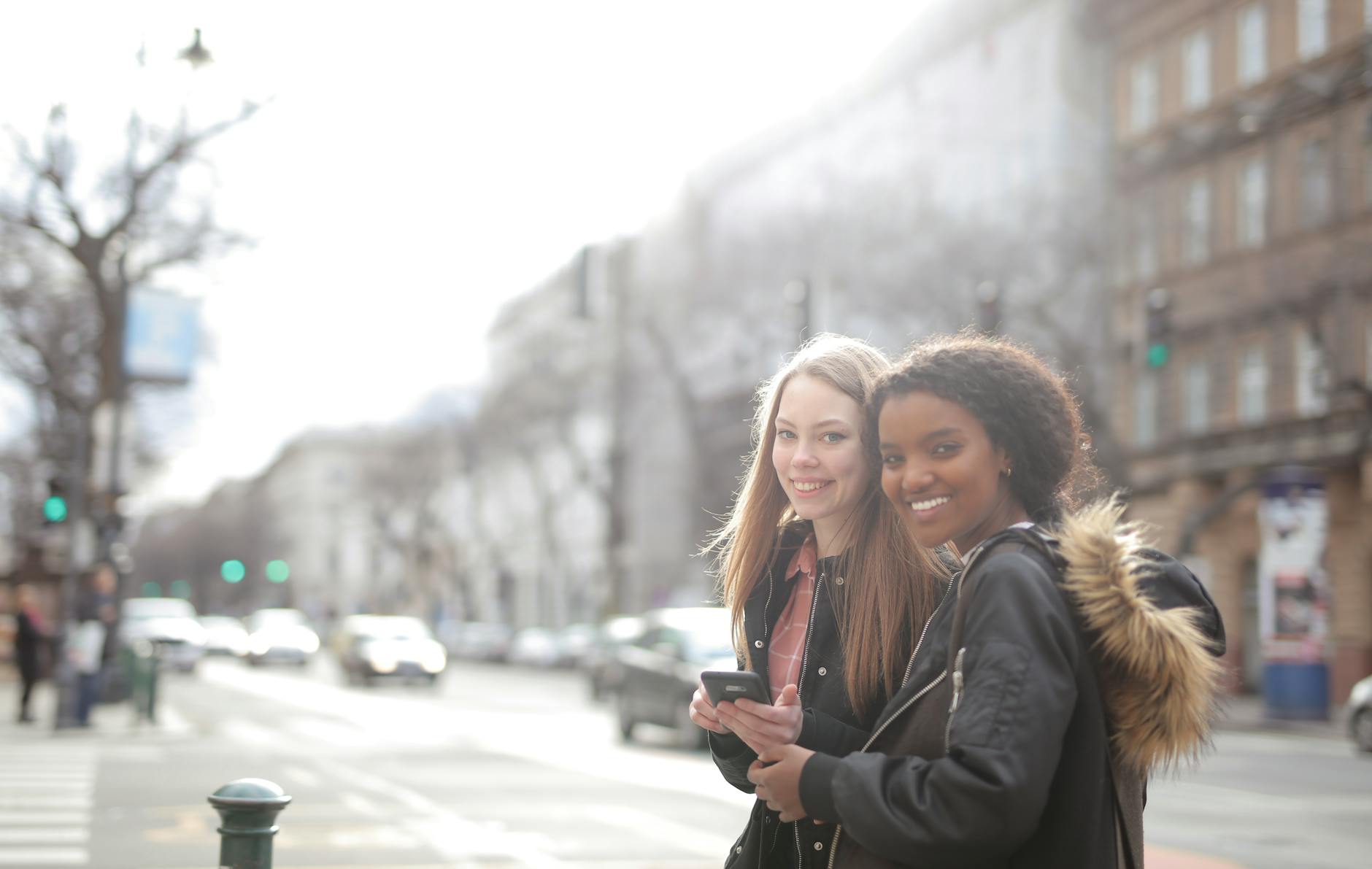 Two young women smiling and holding phones while standing on a bustling city street.