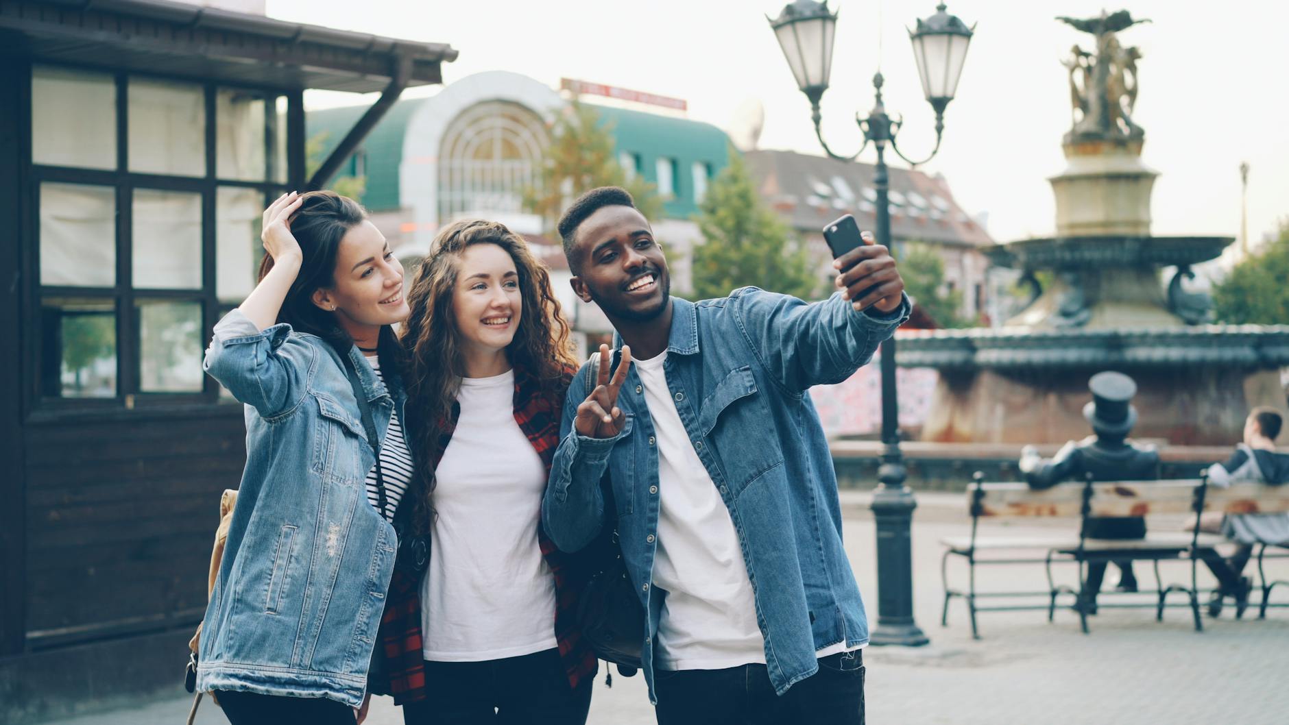 Three diverse friends smiling and taking a selfie near a city fountain during daytime.