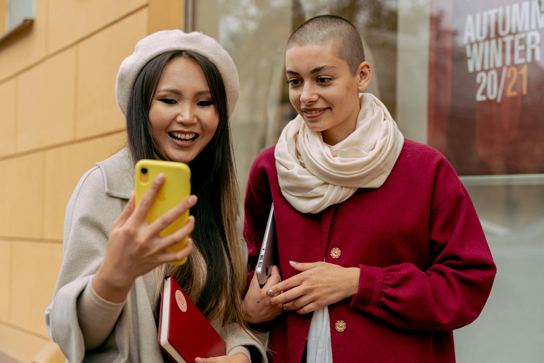 Two fashionable women share a happy moment while looking at a phone outdoors. Autumn vibes.