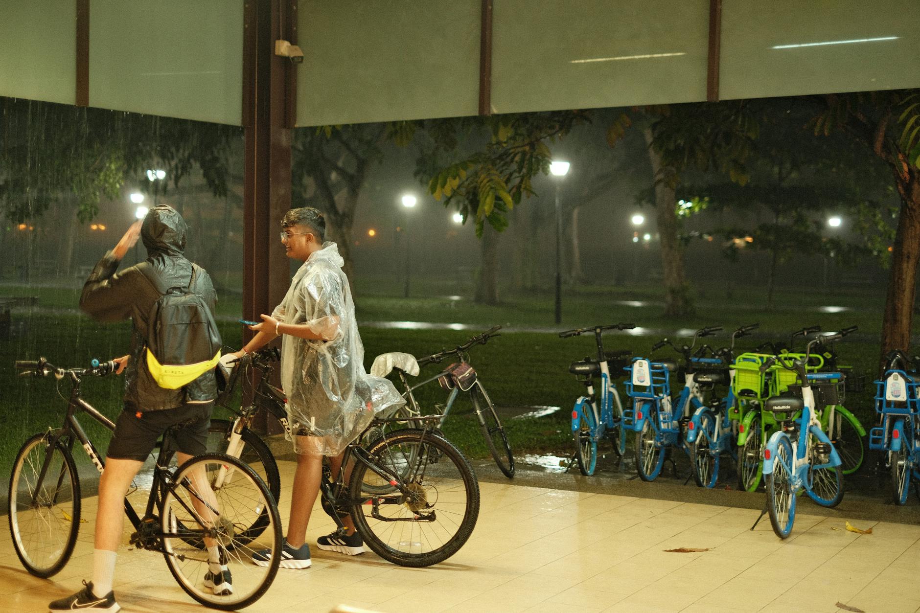 Two cyclists take shelter during a rainy night in a park, surrounded by rental bikes.
