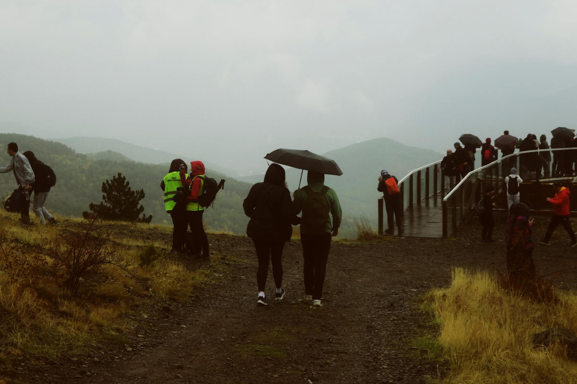 Hikers with umbrellas enjoying a rainy day at a mountain viewpoint with scenic landscape.