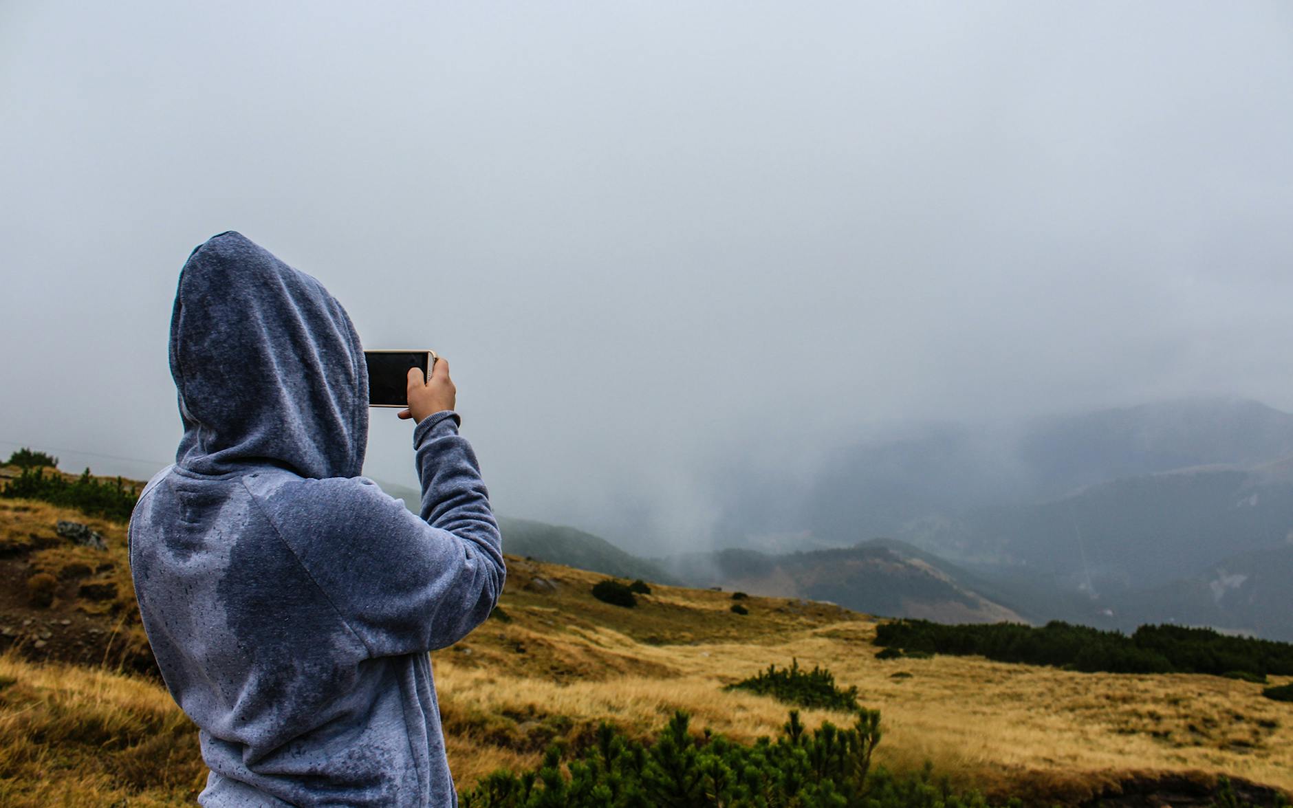 A person takes photos of misty mountains in a hoodie outdoors.