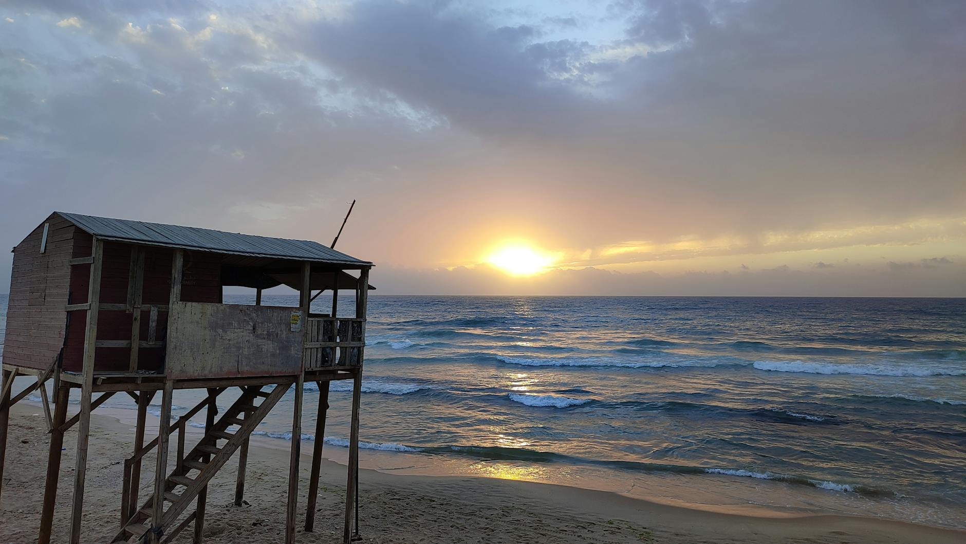 Tranquil sunrise over a sandy beach with a rustic lifeguard hut silhouetted against the calm waves.