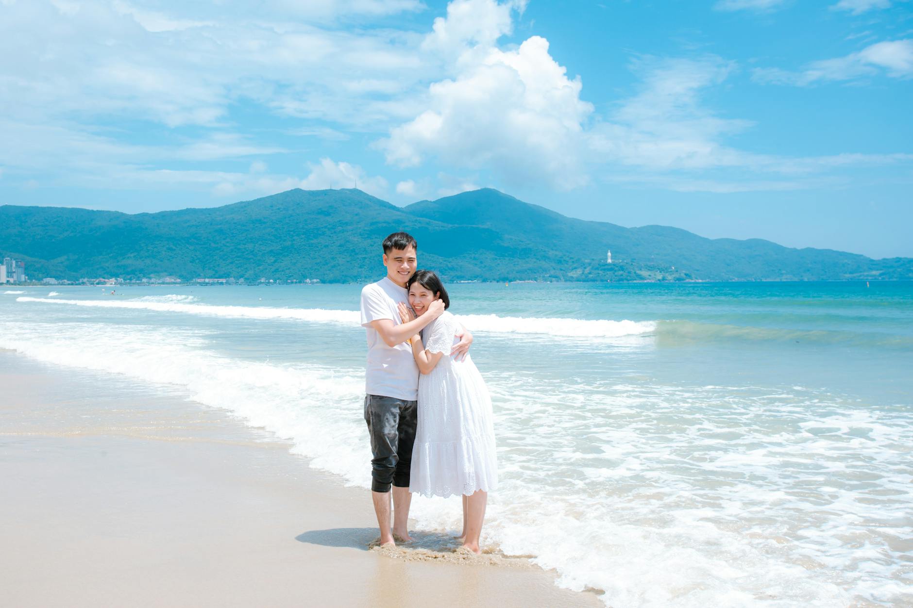 A happy couple embracing on a sunny day at Da Nang beach in Vietnam with mountains in the background.