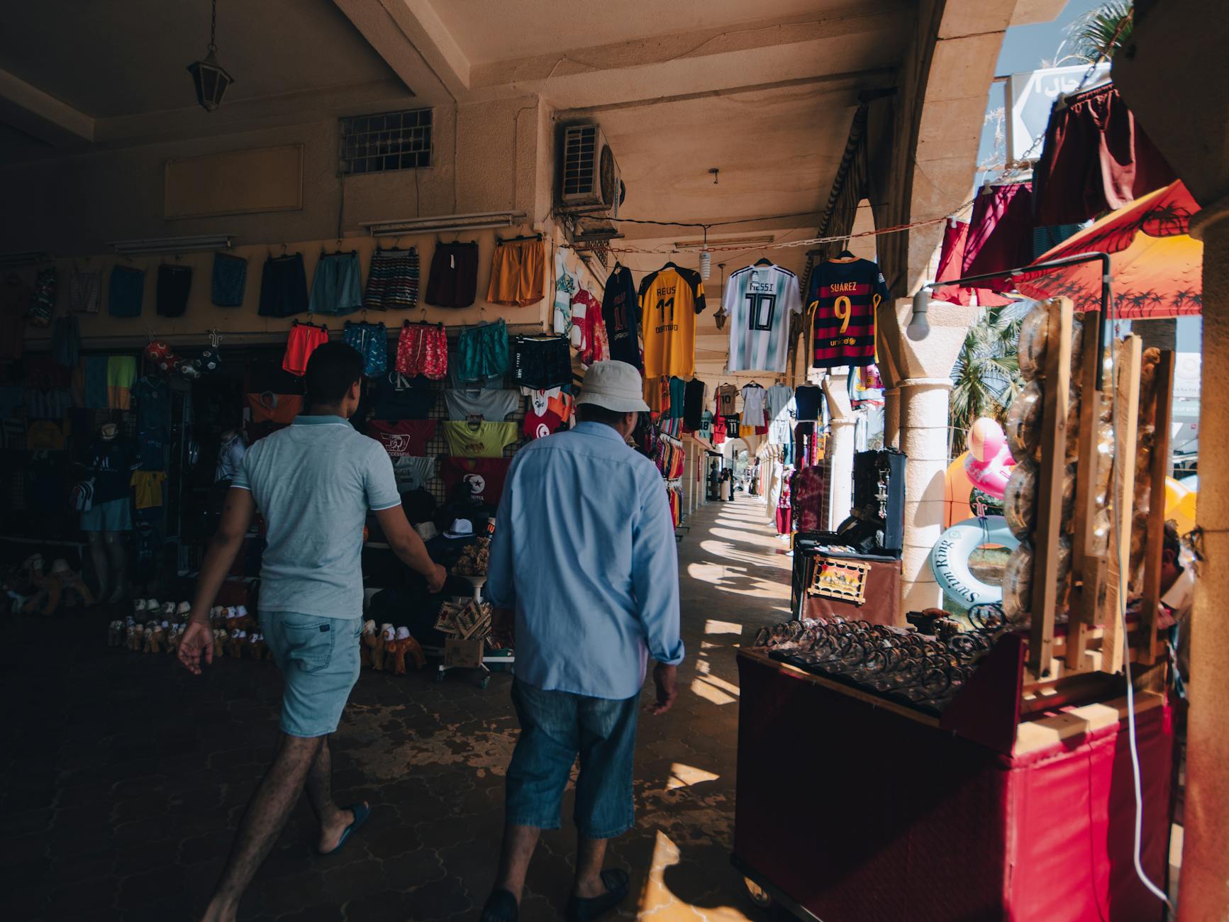 Two men walk through a vibrant clothing market under the building's arched hallway.